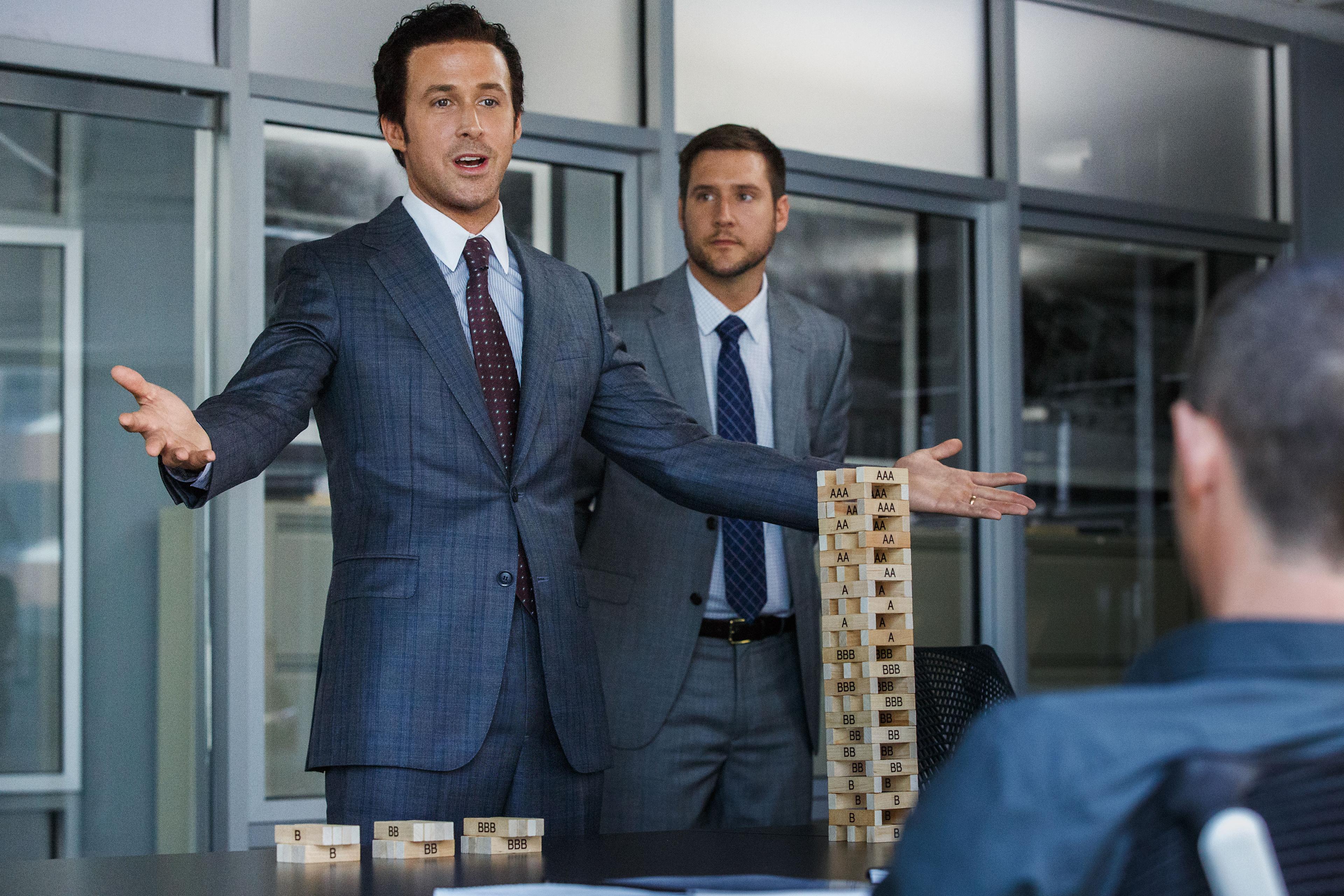 Photo of a businessman in a suit explaining with a Jenga tower, another man stands behind him, both in an office setting.