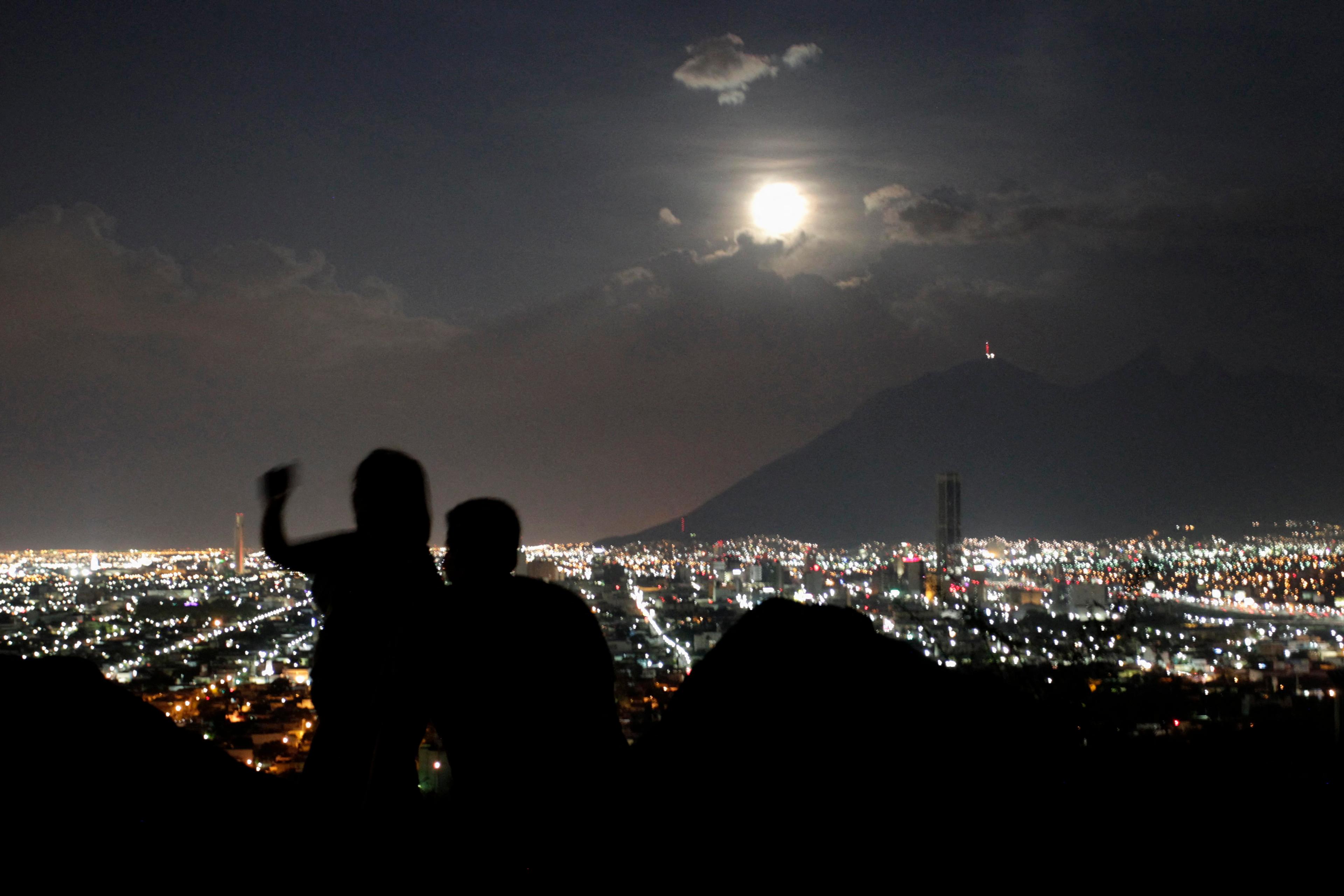 A cityscape at night with two silhouetted figures in the foreground and a full moon above a mountain in the background.