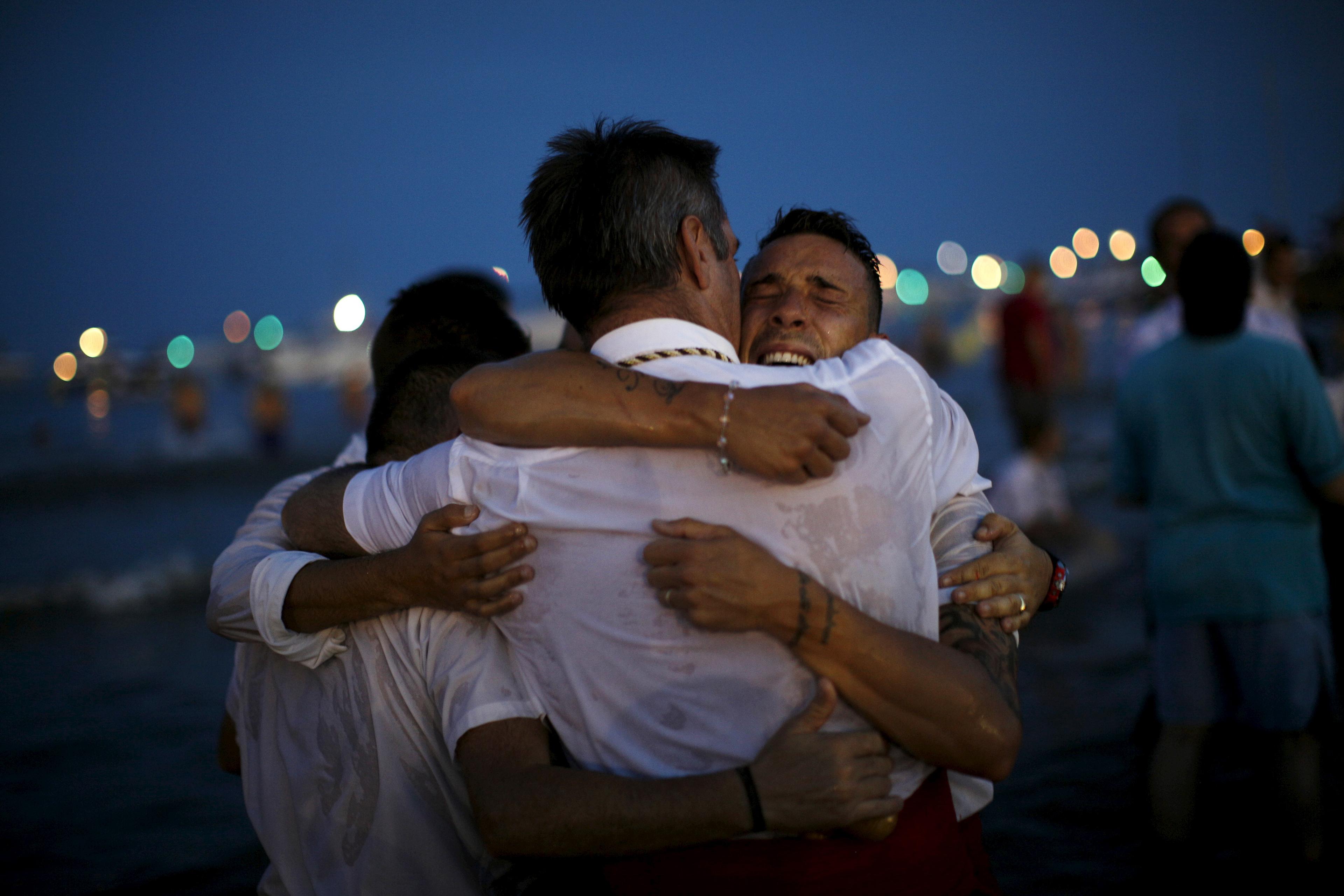 Photo of four men embracing on a beach at night with colourful lights blurred in the background.