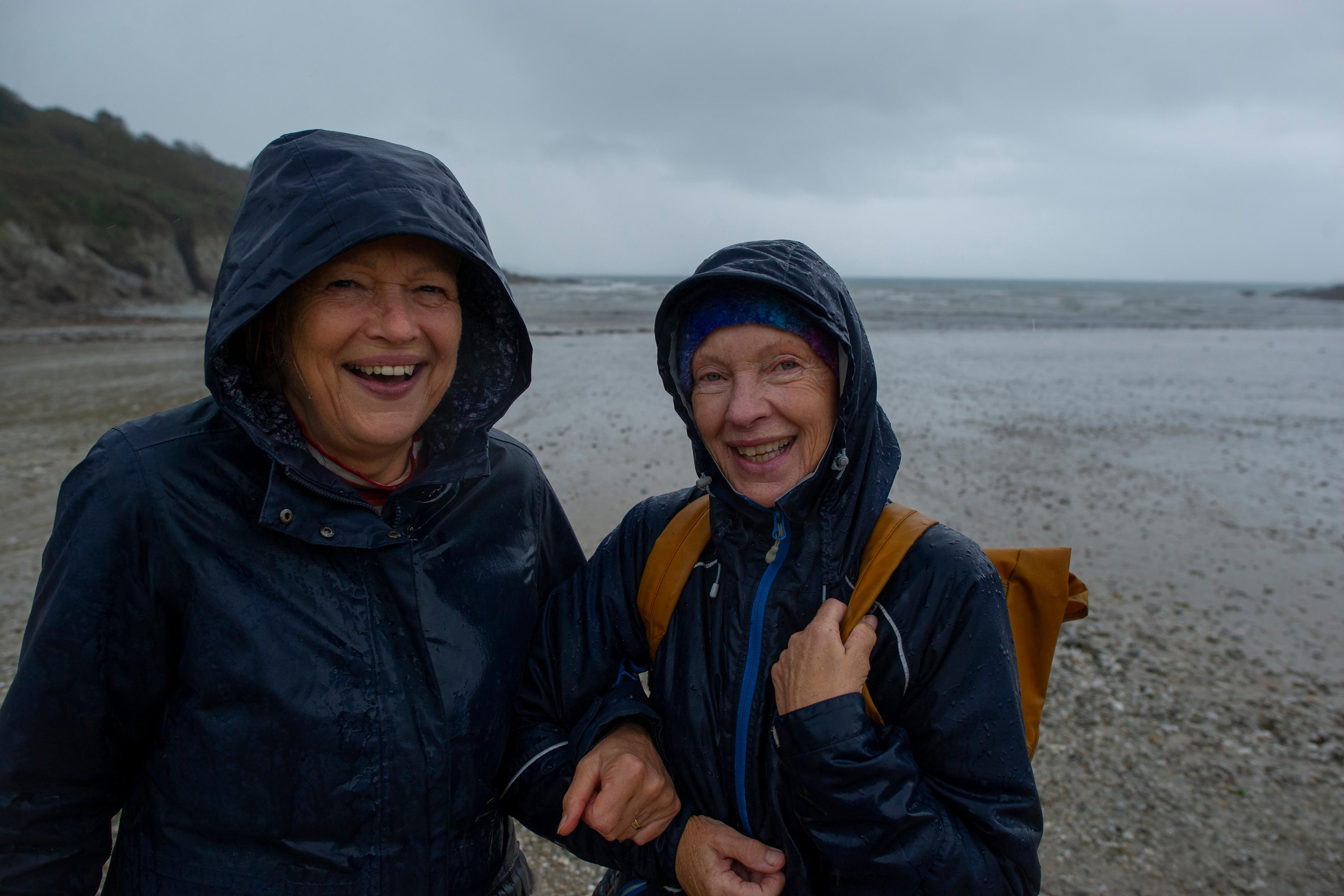 Photo of two smiling people in waterproof jackets on a rainy beach, grey skies in the background.