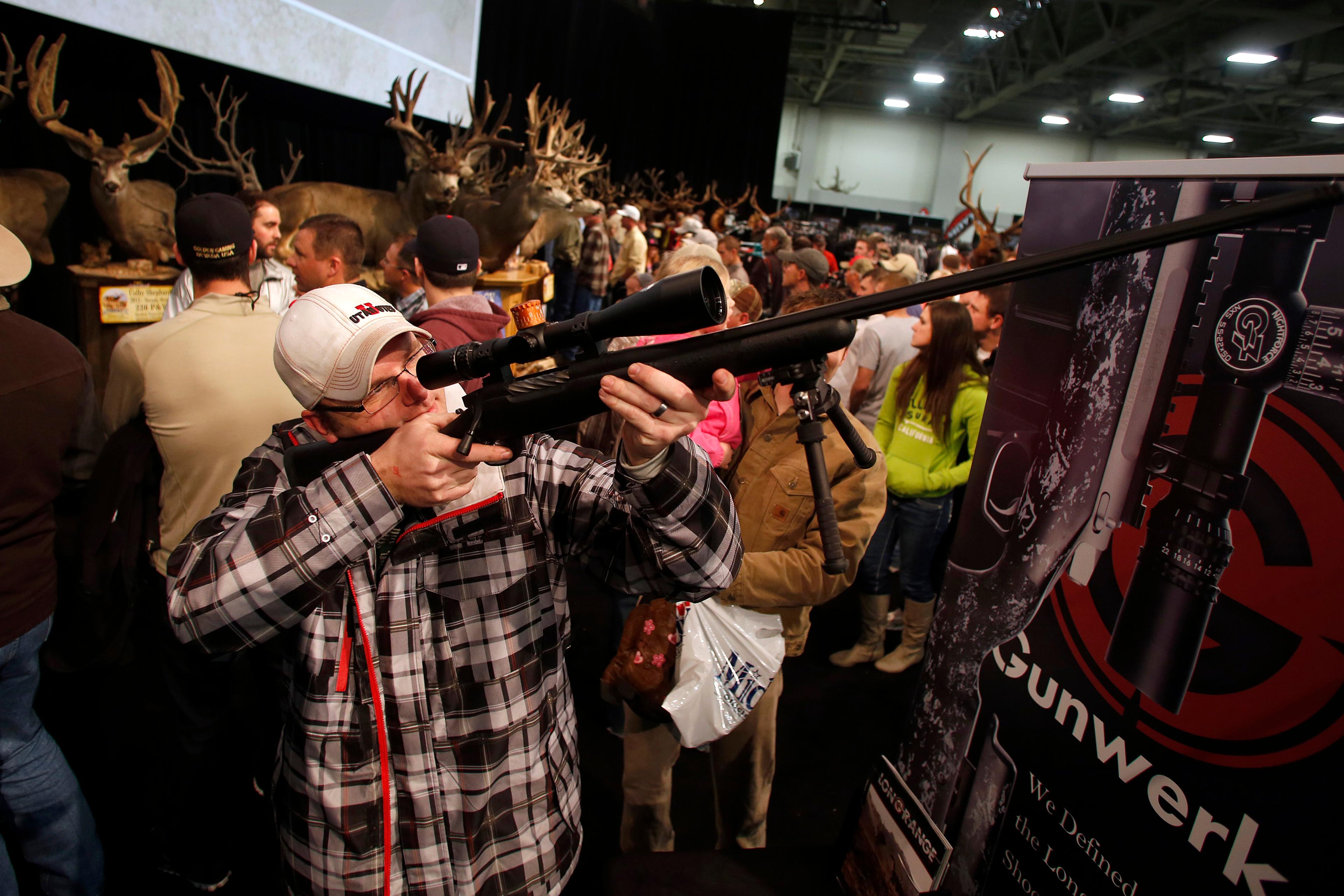 A man looking through the scope of a rifle in a crowded exhibition with mounted deer heads in the background.
