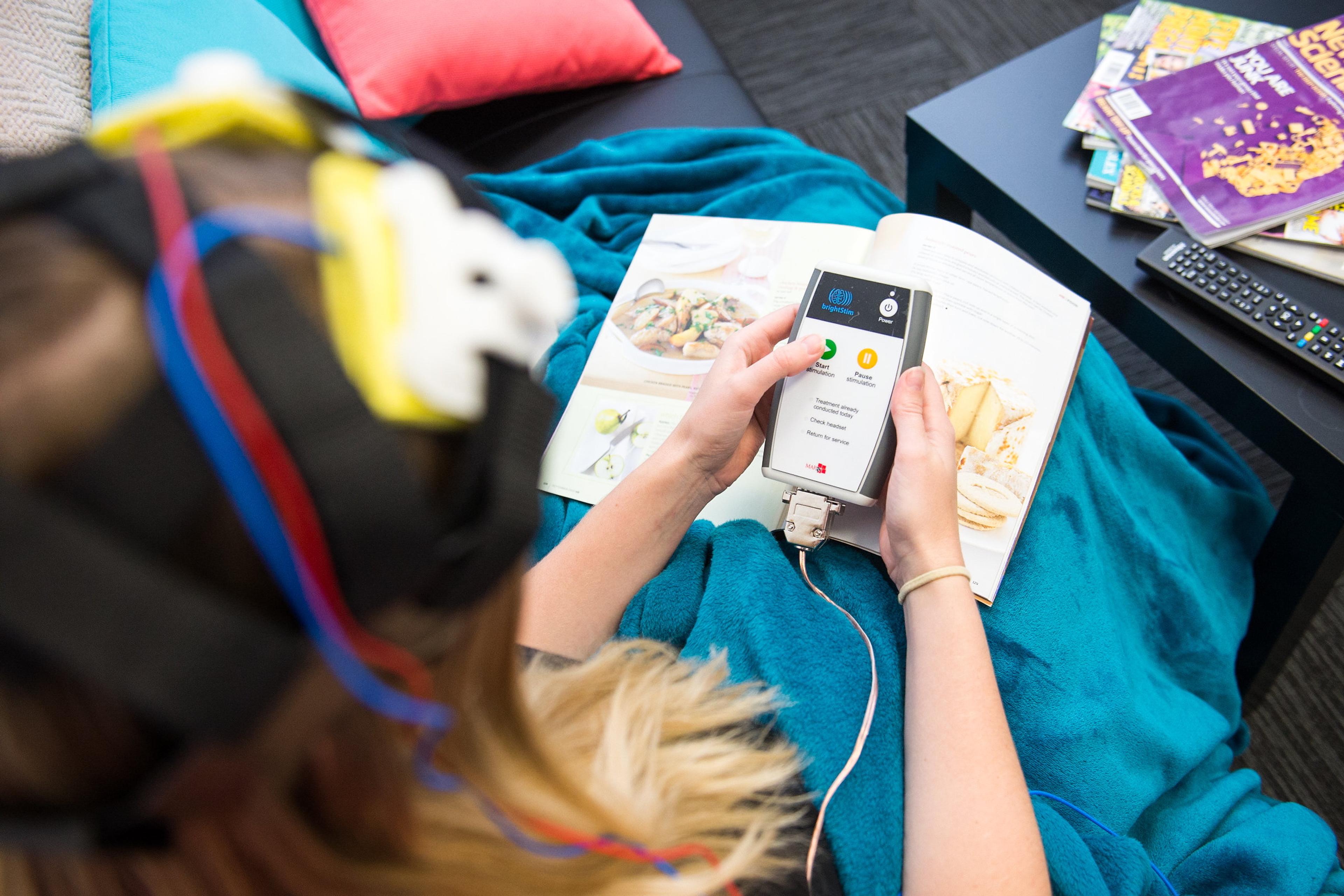 Photo of a person with a headset holding a device, looking at a cookbook on a sofa with magazines and a remote nearby.