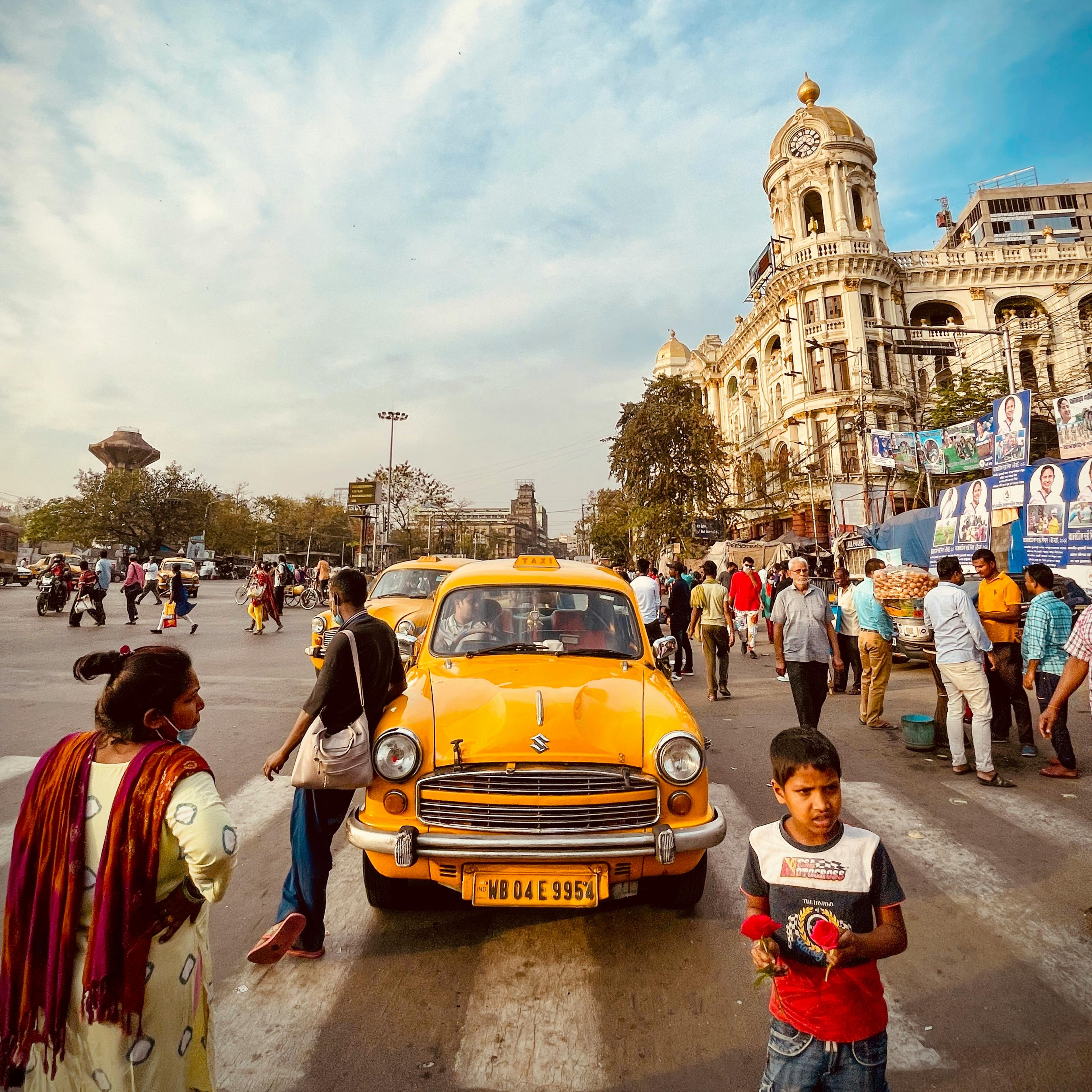 A yellow taxi on a busy street, people walking, child holding flowers, historic building in the background under a blue sky.