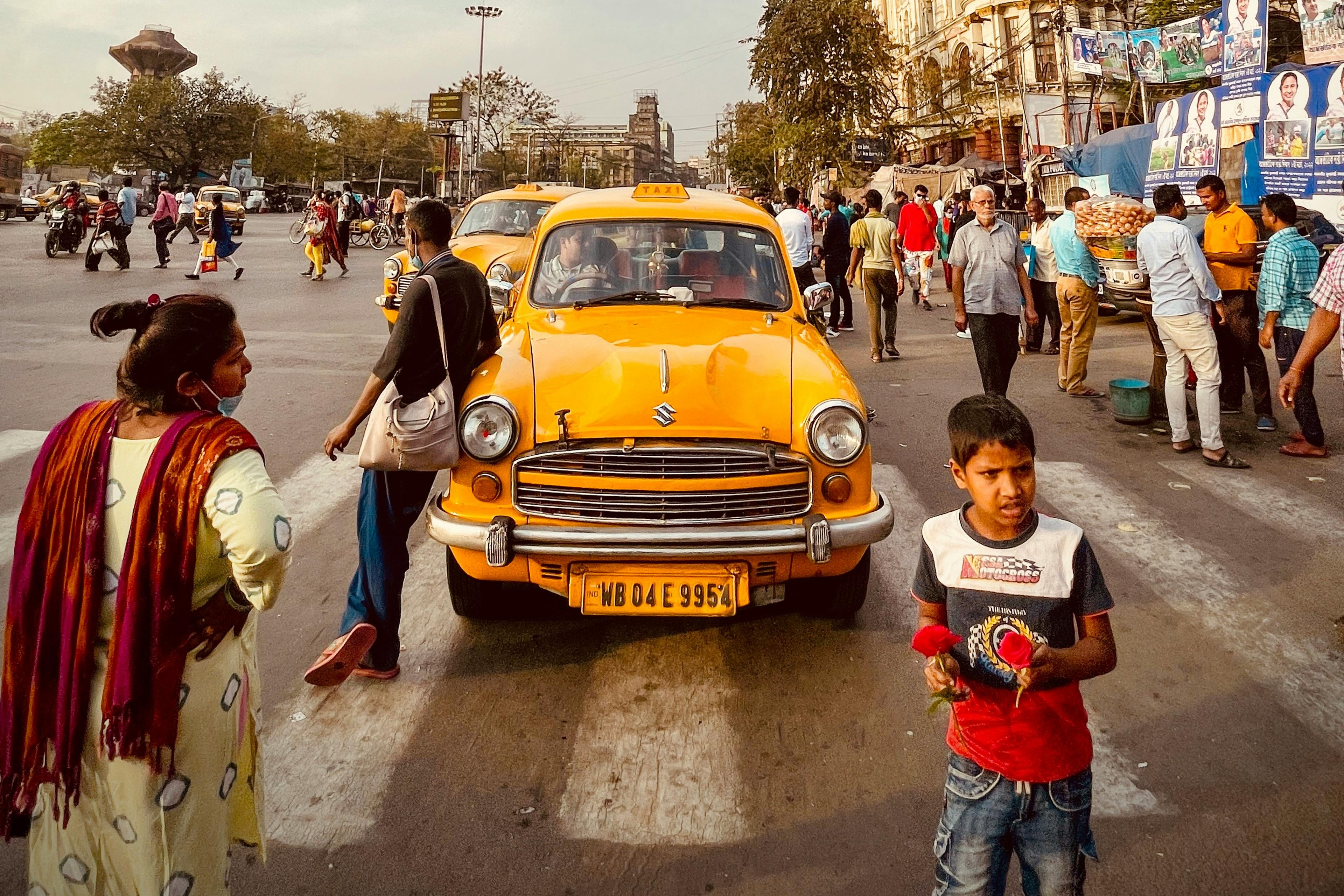 A yellow taxi on a busy street, people walking, child holding flowers, historic building in the background under a blue sky.