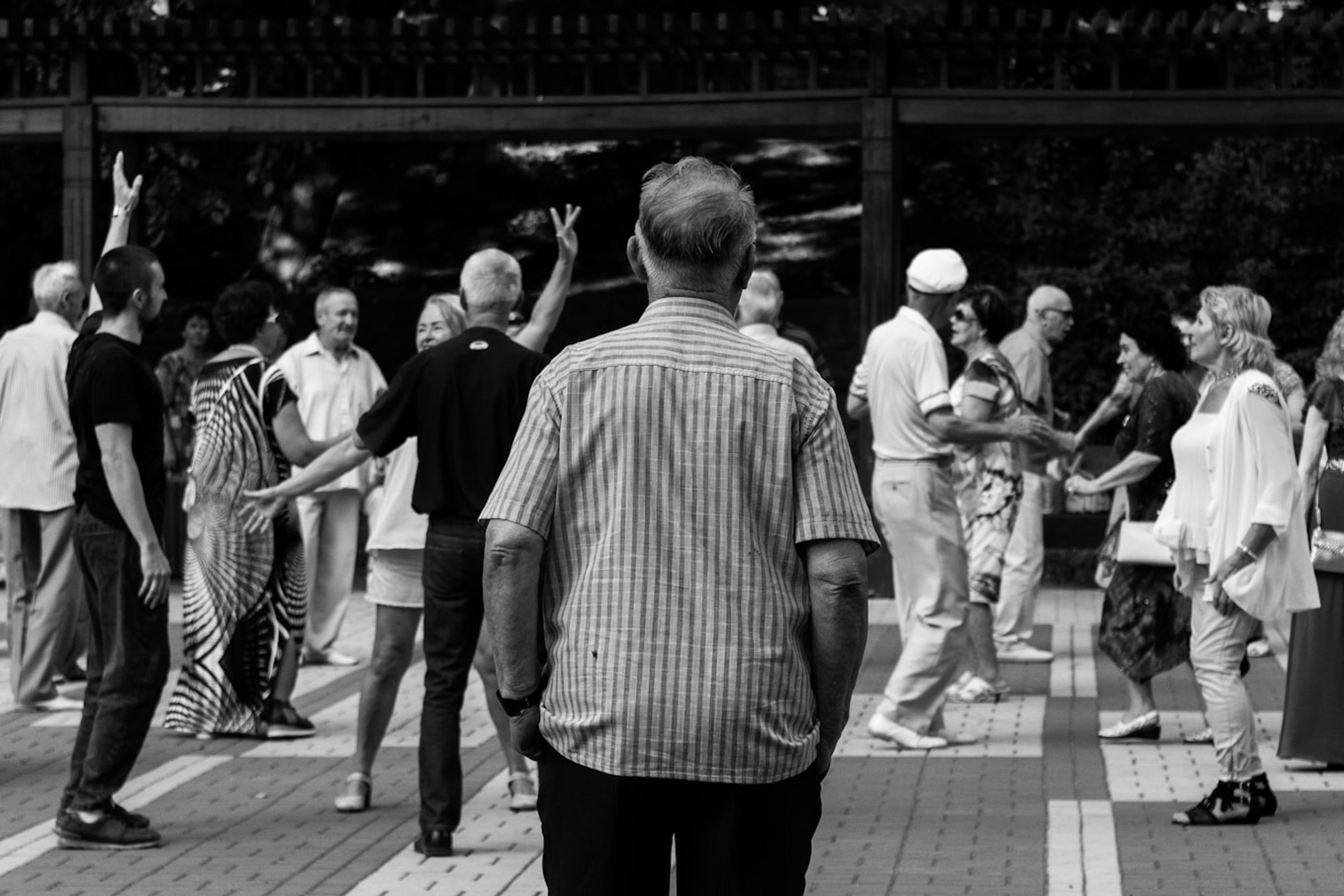 Black and white photo of a group of people dancing outdoors, with a man in a striped shirt in the foreground.