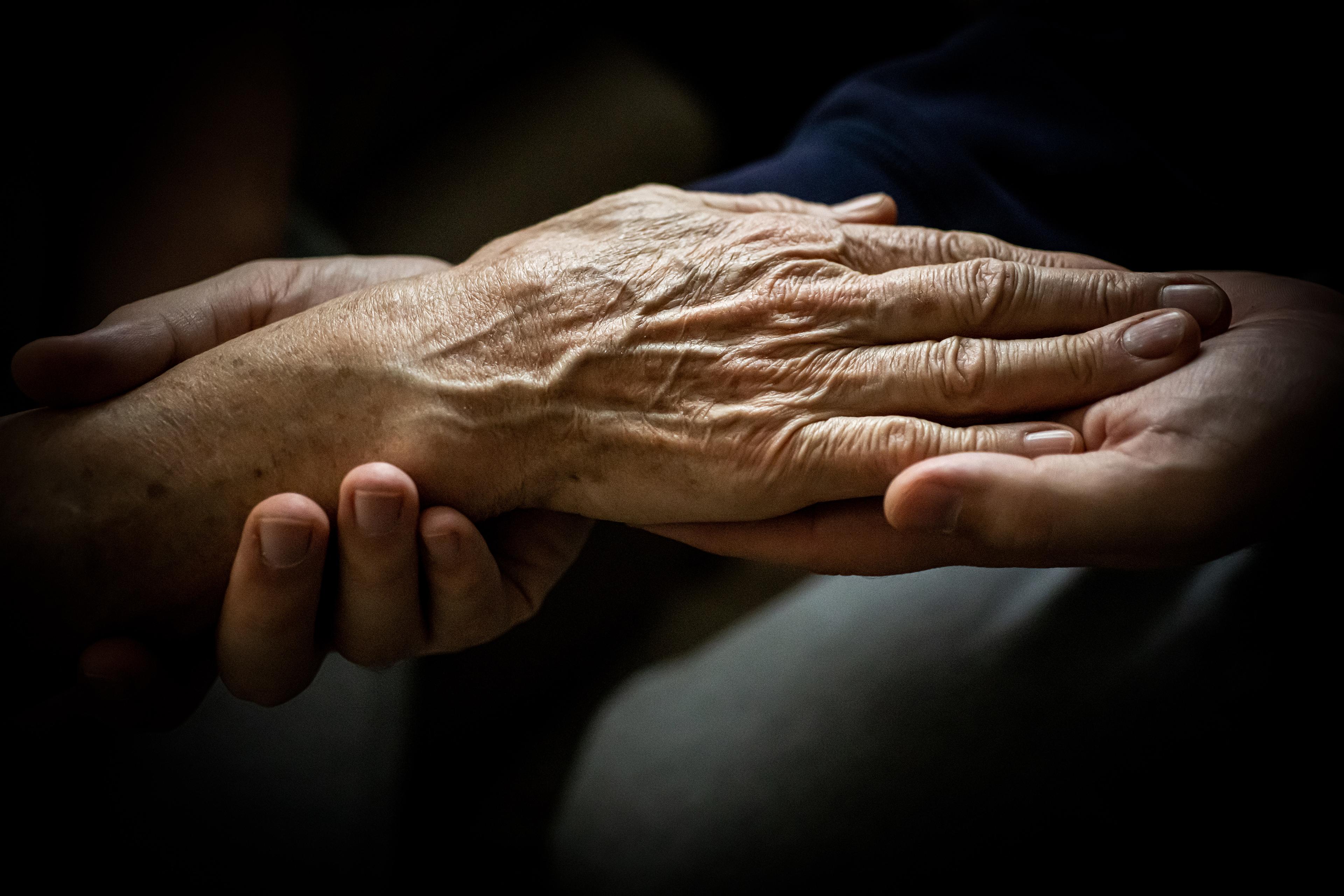 An elderly hand resting being held by younger hands against a dark background.
