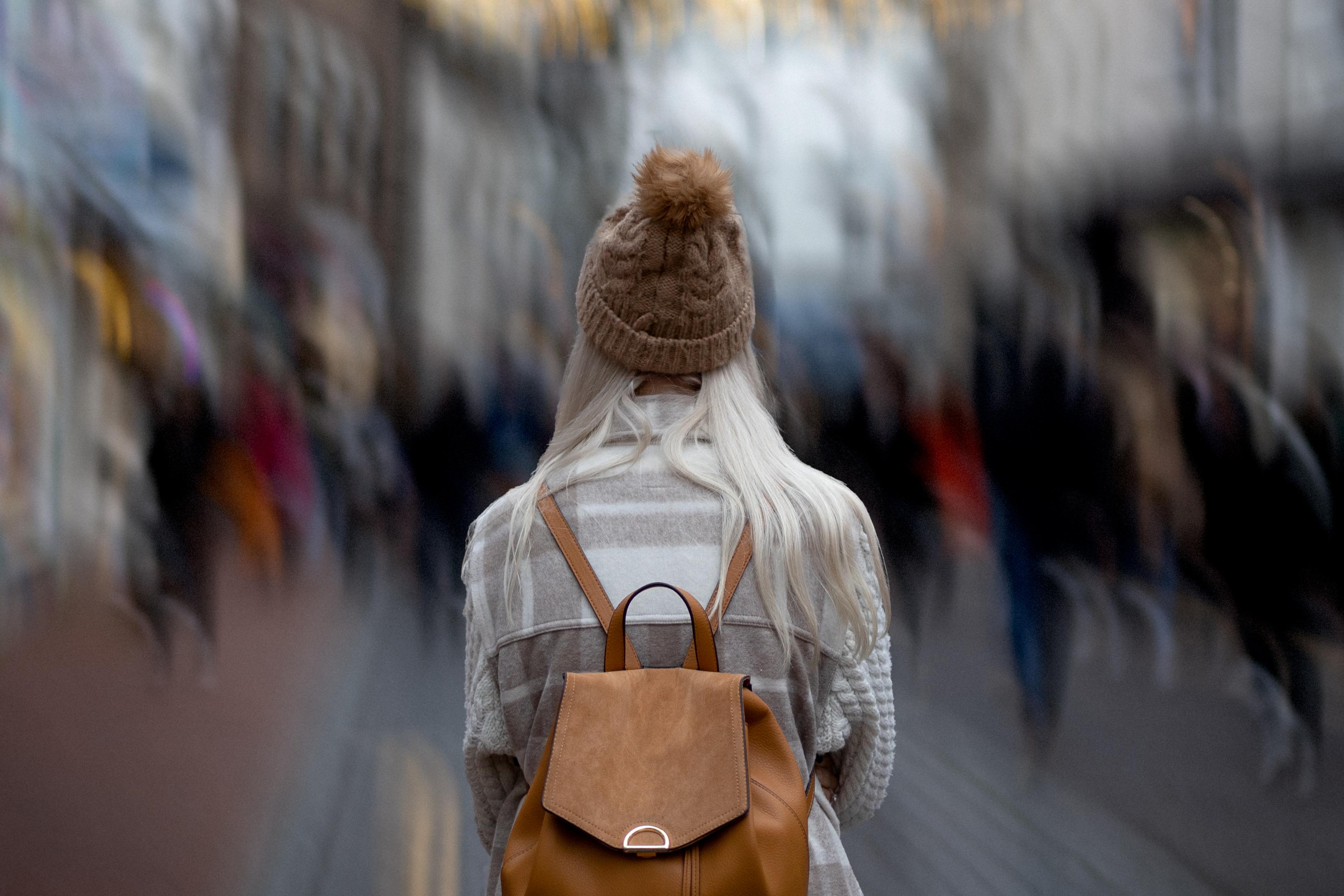 A person with long blonde hair in a beanie and coat, facing away from the camera, wearing a brown backpack in a blurred busy street.