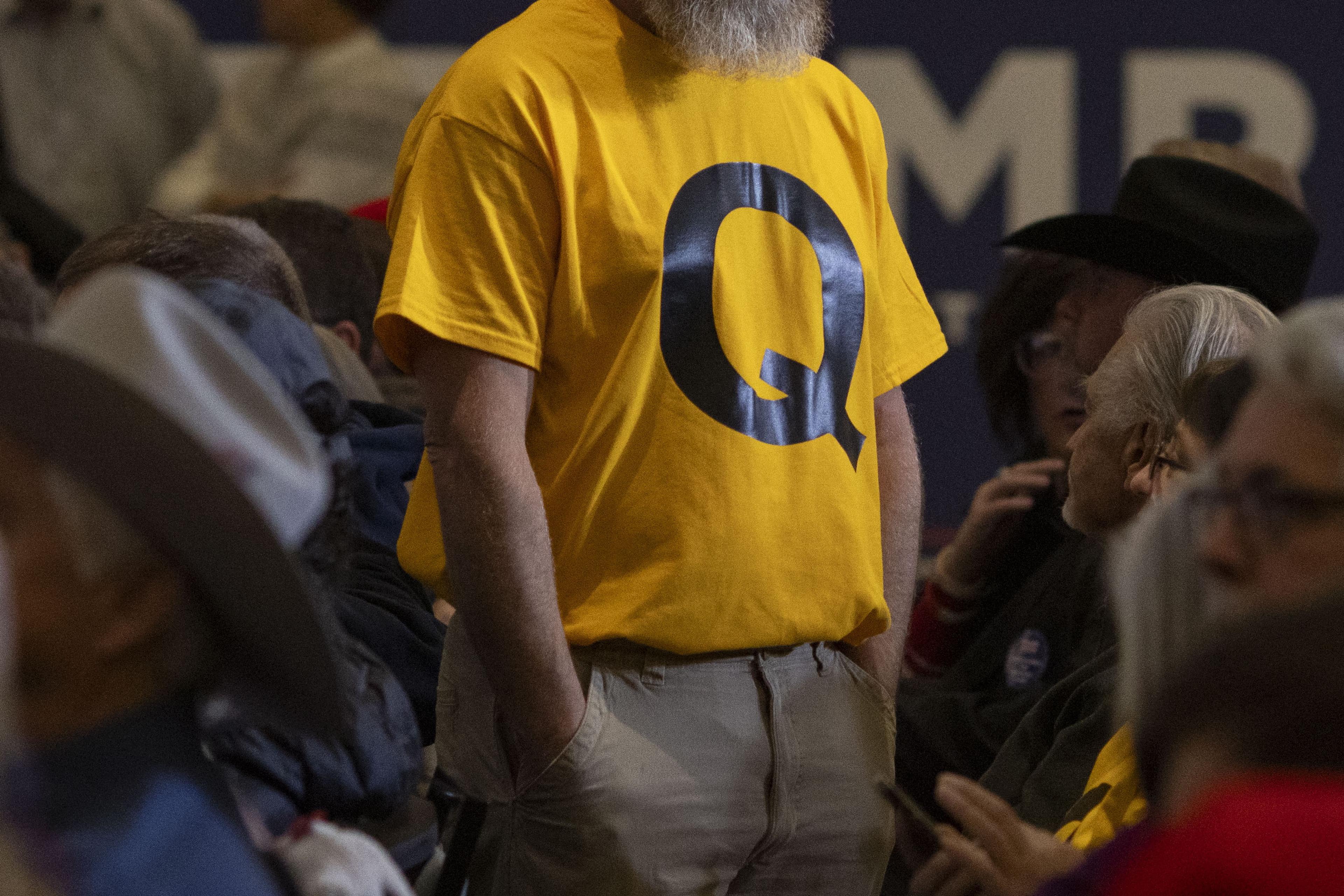 Photo of a person in a yellow T-shirt with a large black letter Q standing among seated people at an indoor event.