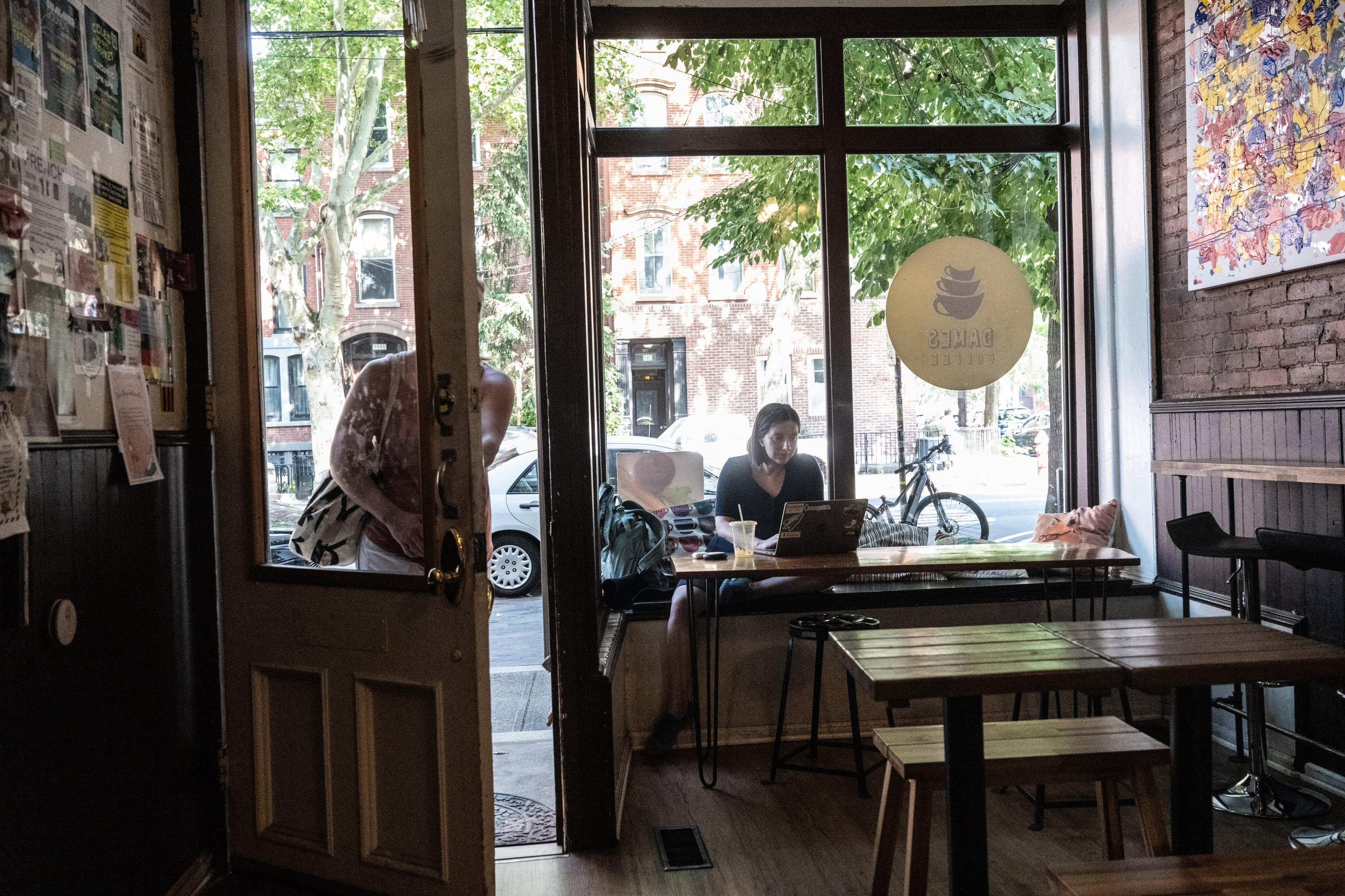 Photo of a cafe interior with a person working on a laptop by a window, open door and street view with bicycle outside.