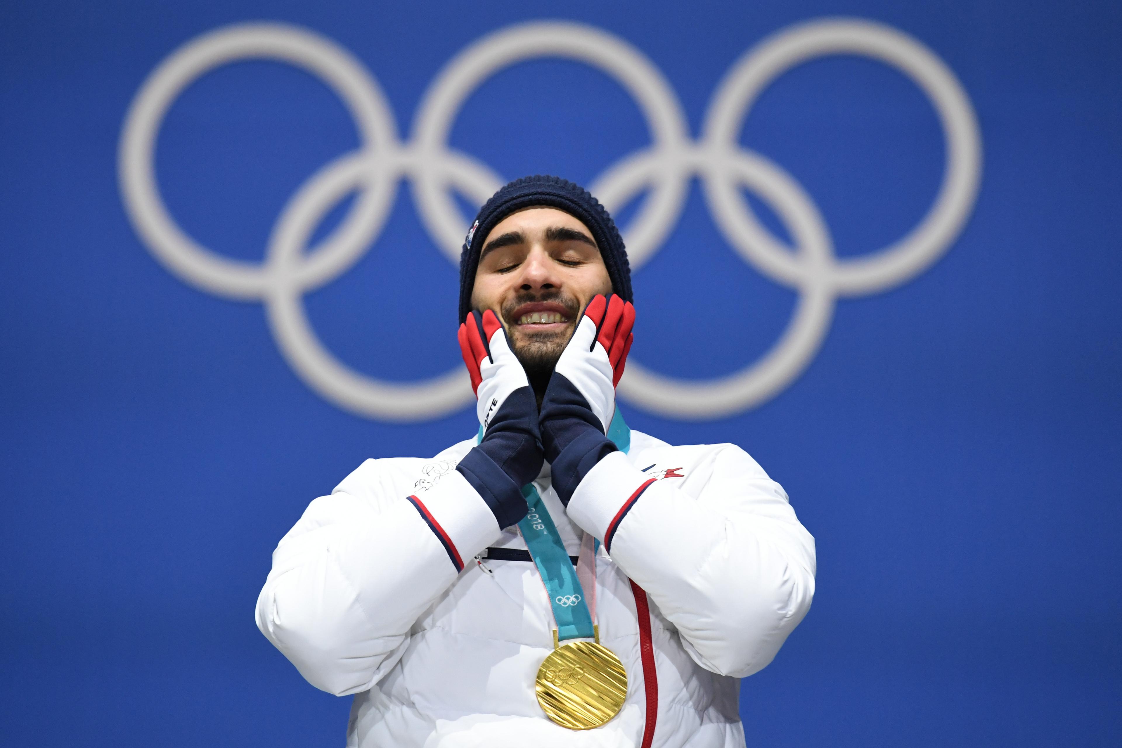 Photo of an athlete in a white jacket with a gold medal, smiling with hands on face against an Olympic rings backdrop.