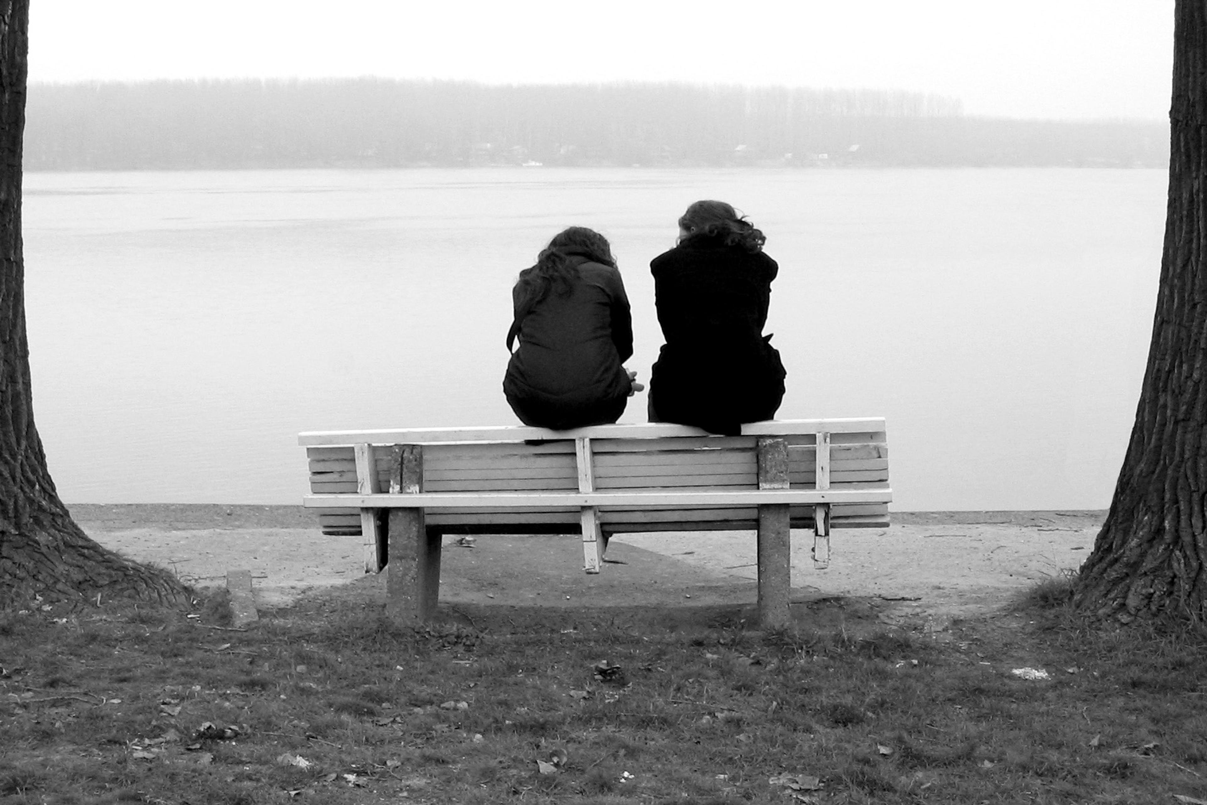Black and white photo of two people sitting on a bench by a lake between two trees.