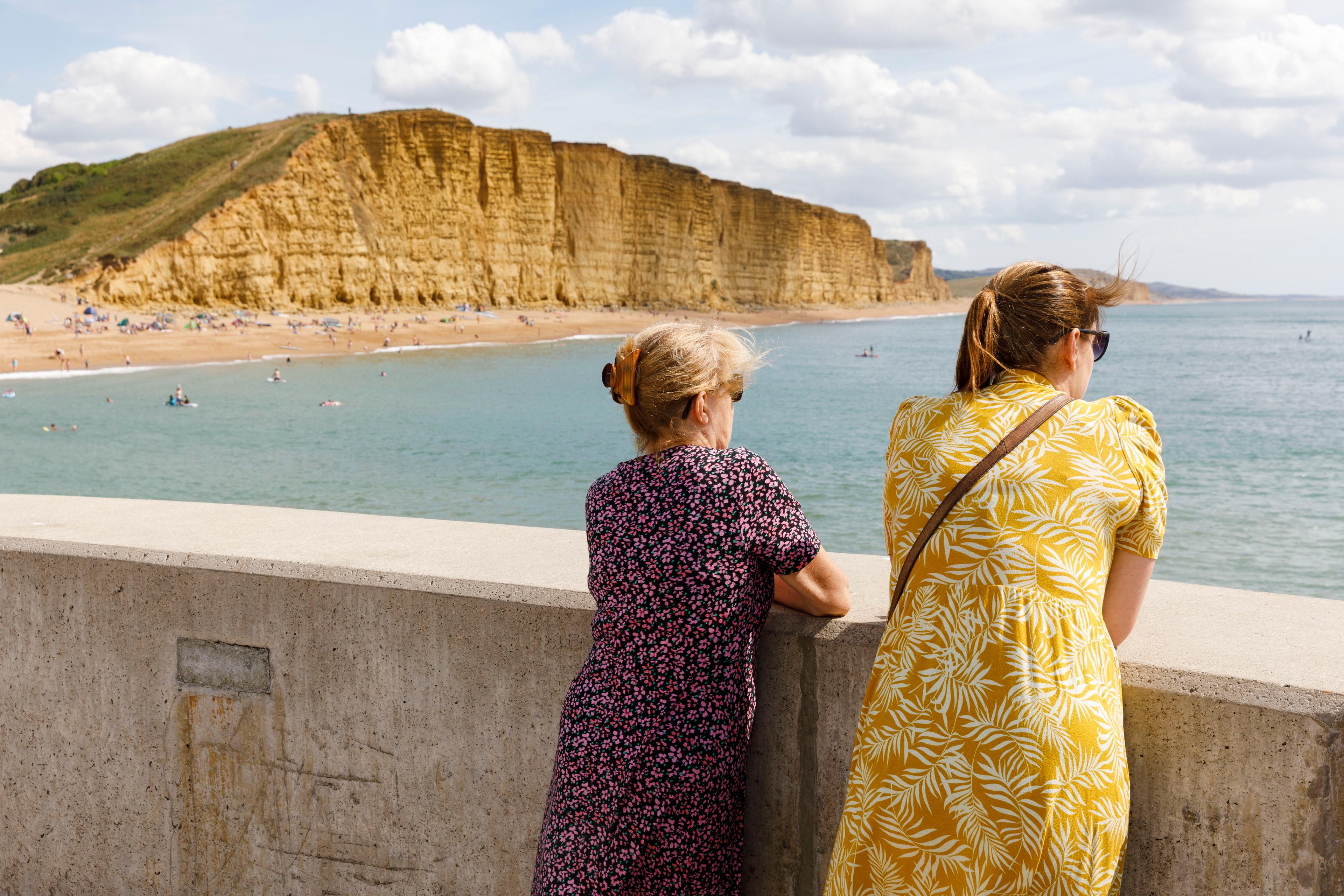 Photo of two women leaning on a concrete wall overlooking a sandy beach and cliffs by a calm blue sea.