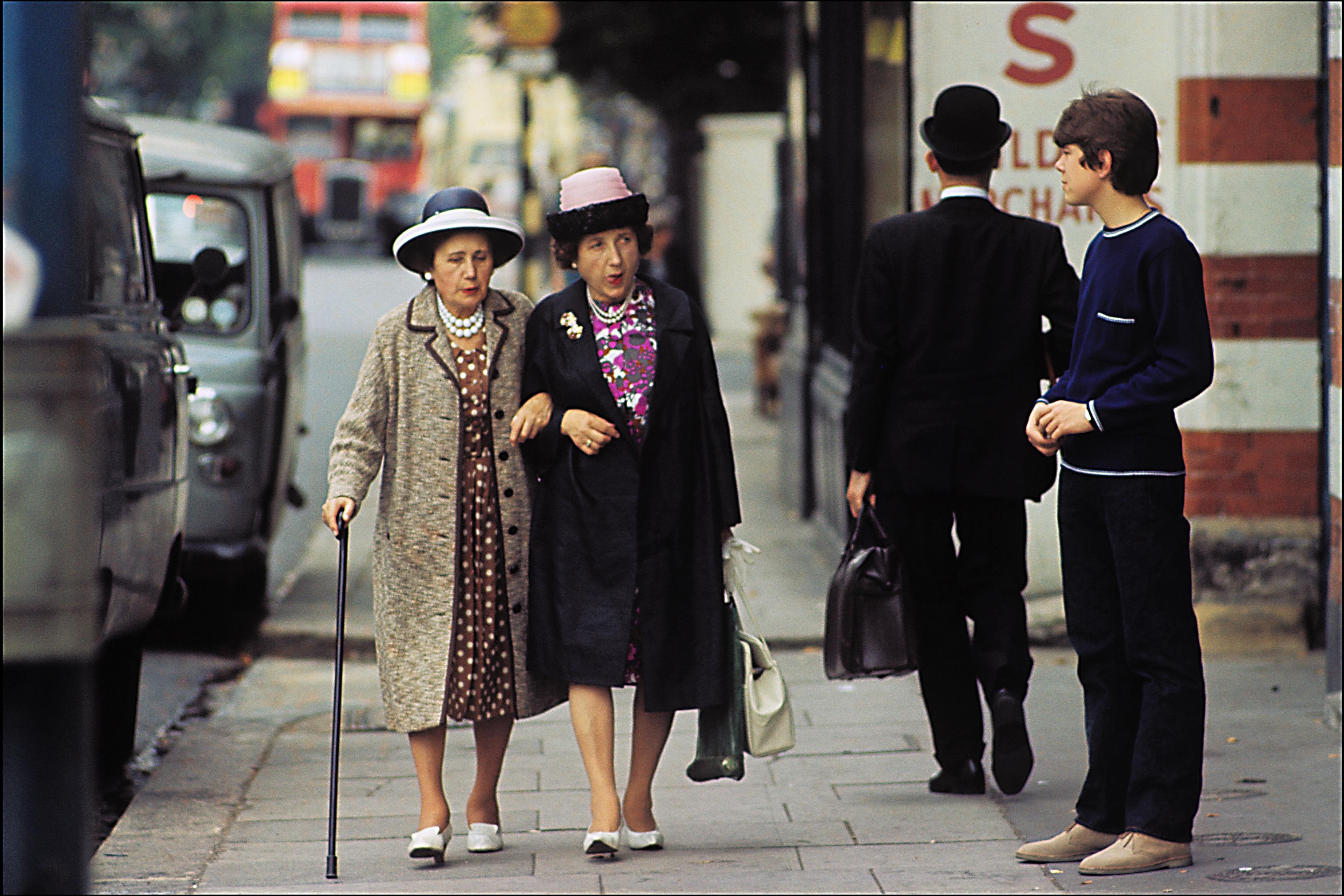 Photo of two elderly women walking together on a city pavement, with a red bus and two other people in the background.