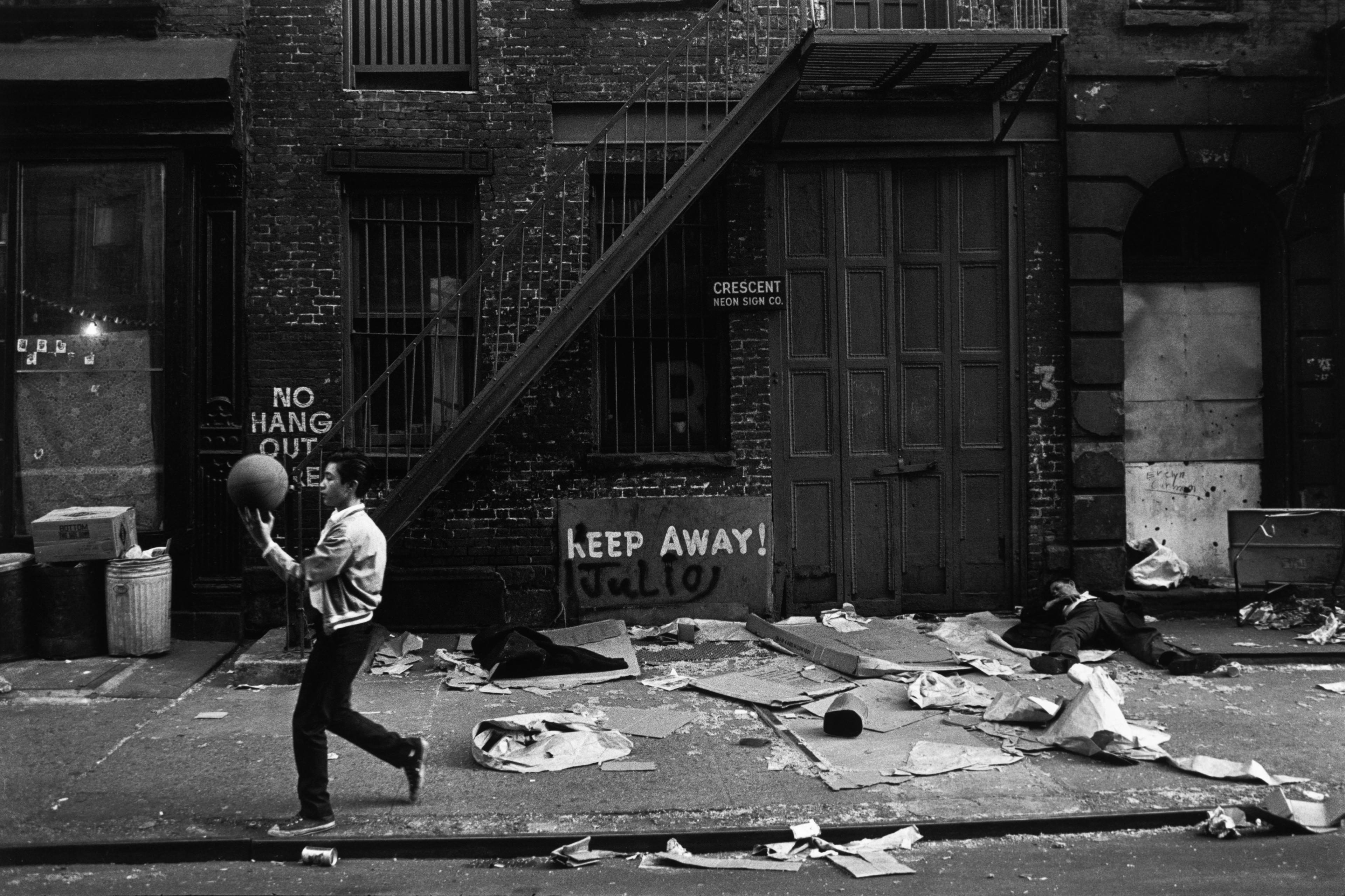 Black and white photo of a man with a ball on a littered street, graffiti and a fire escape visible on a brick building.