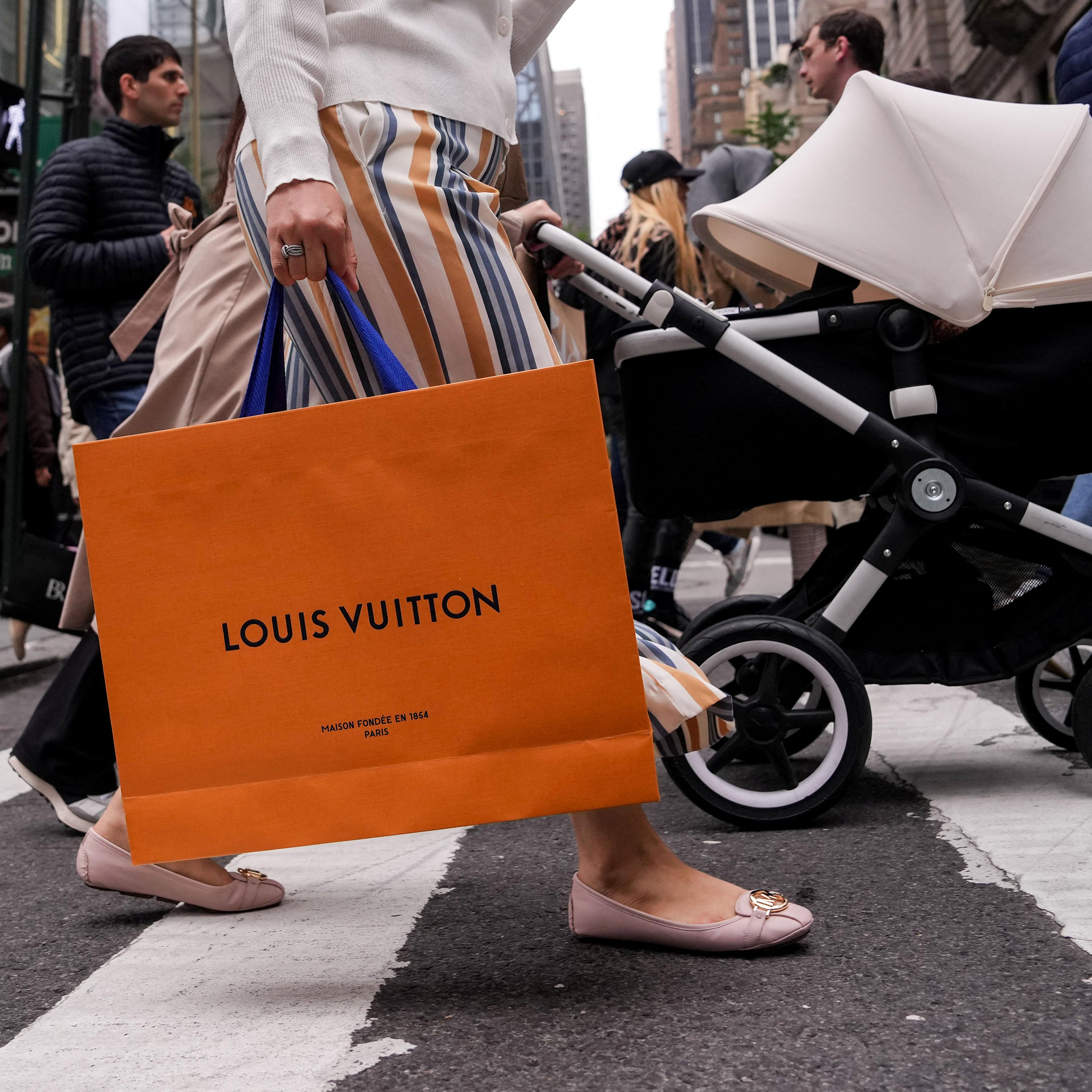 A person crossing a busy city street holding a Louis Vuitton bag with other pedestrians in the background.