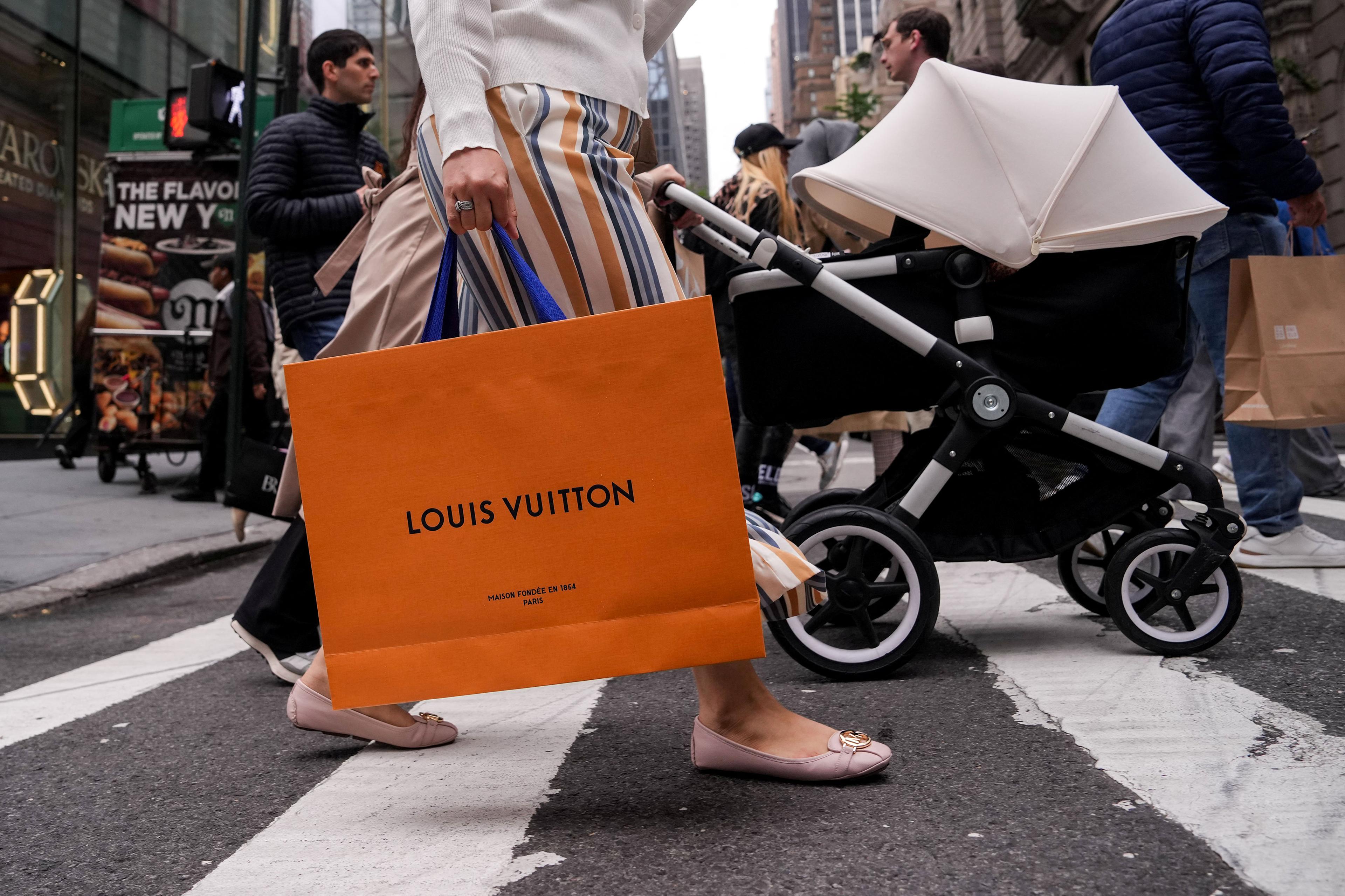 A person crossing a busy city street holding a Louis Vuitton bag with other pedestrians in the background.
