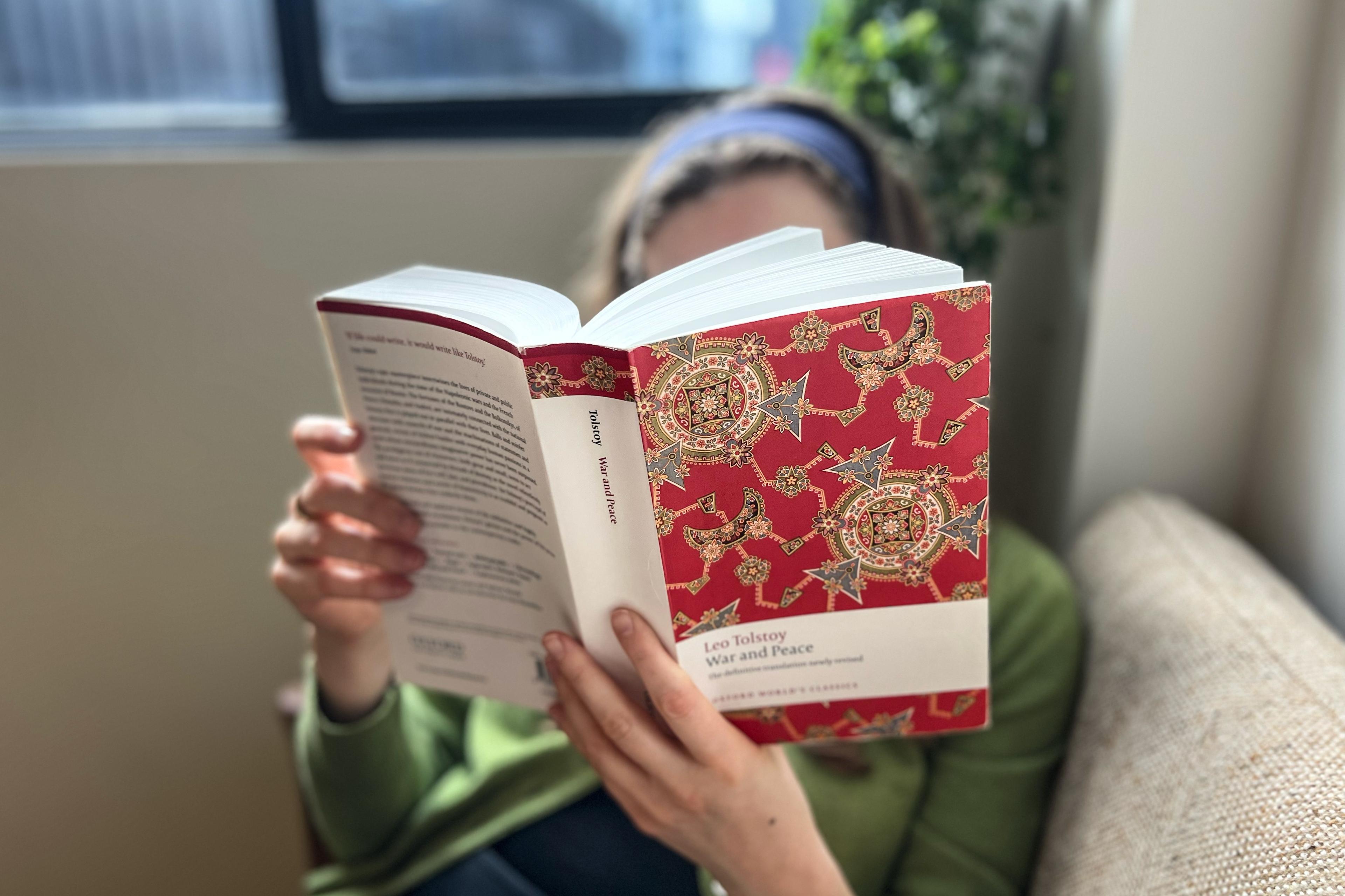 Photo of a person reading Tolstoy’s “War and Peace” with a red decorative cover while sitting near a window.