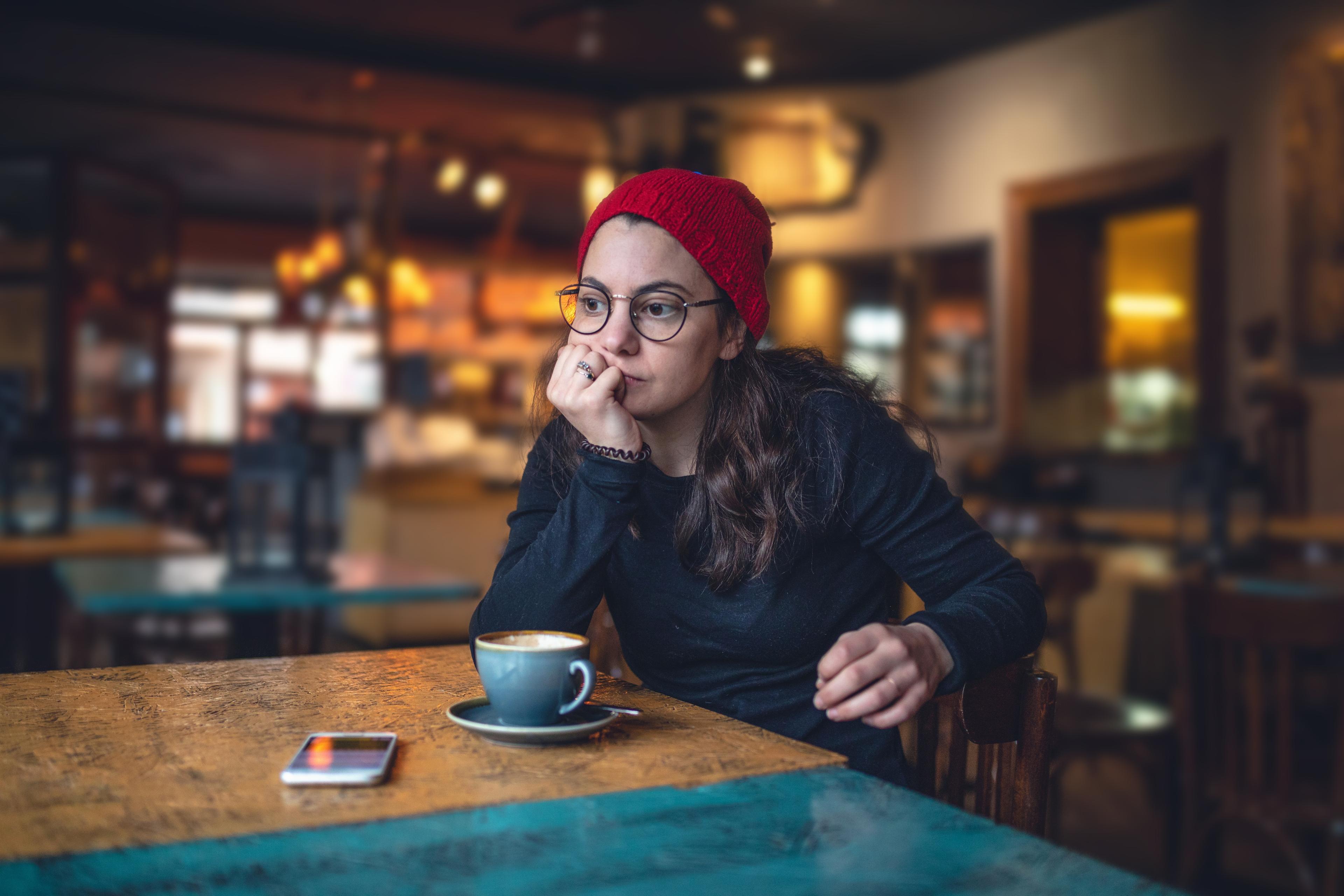Photo of a person in a red hat and glasses sitting thoughtfully in a cafe with a coffee and a phone on the table.
