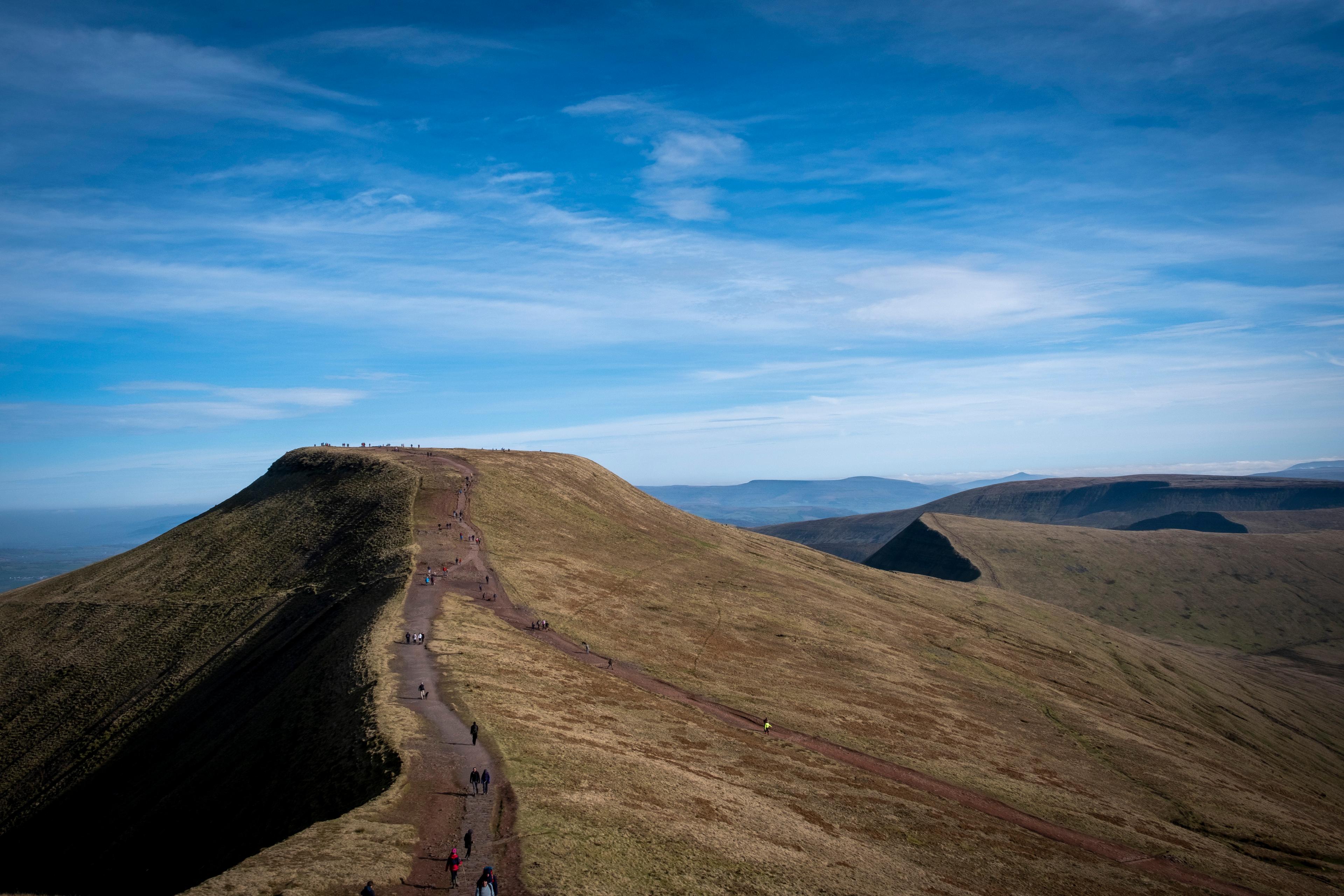 A mountain peak with a walking path and hikers, under a clear blue sky with scattered clouds.