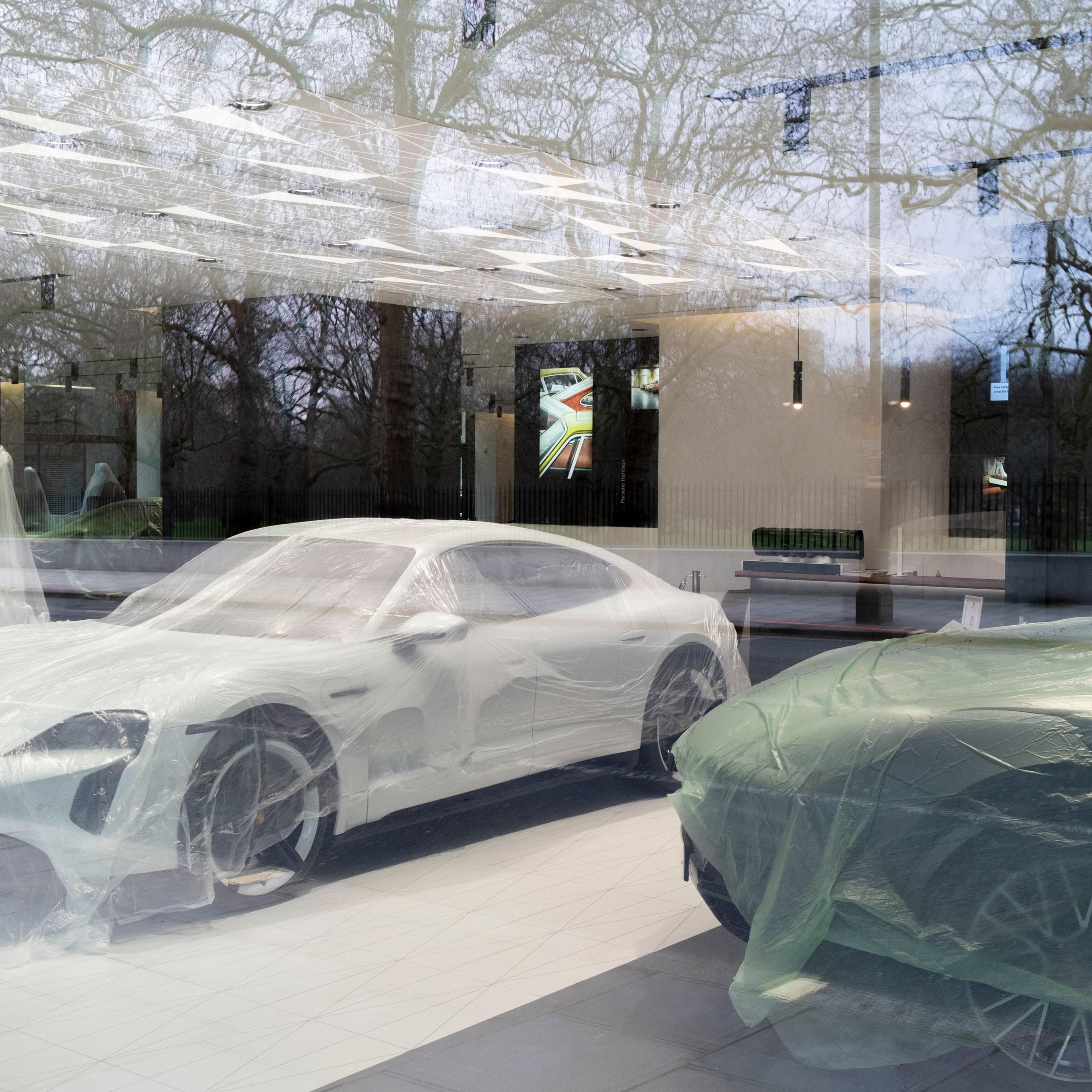 Photo of cars covered in protective plastic inside a showroom with reflections of bare trees in the glass facade.