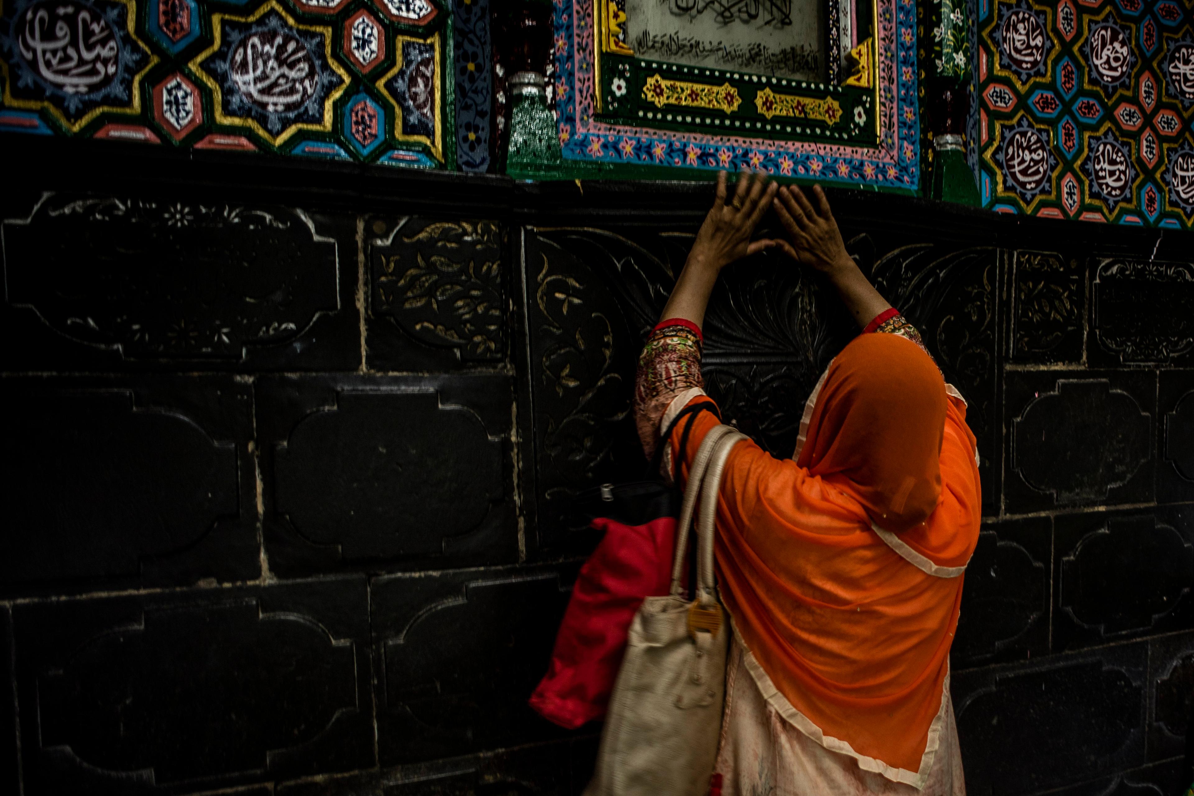 Photo of a woman in an orange shawl touching a decorative wall with colourful, intricate patterns and Islamic calligraphy.