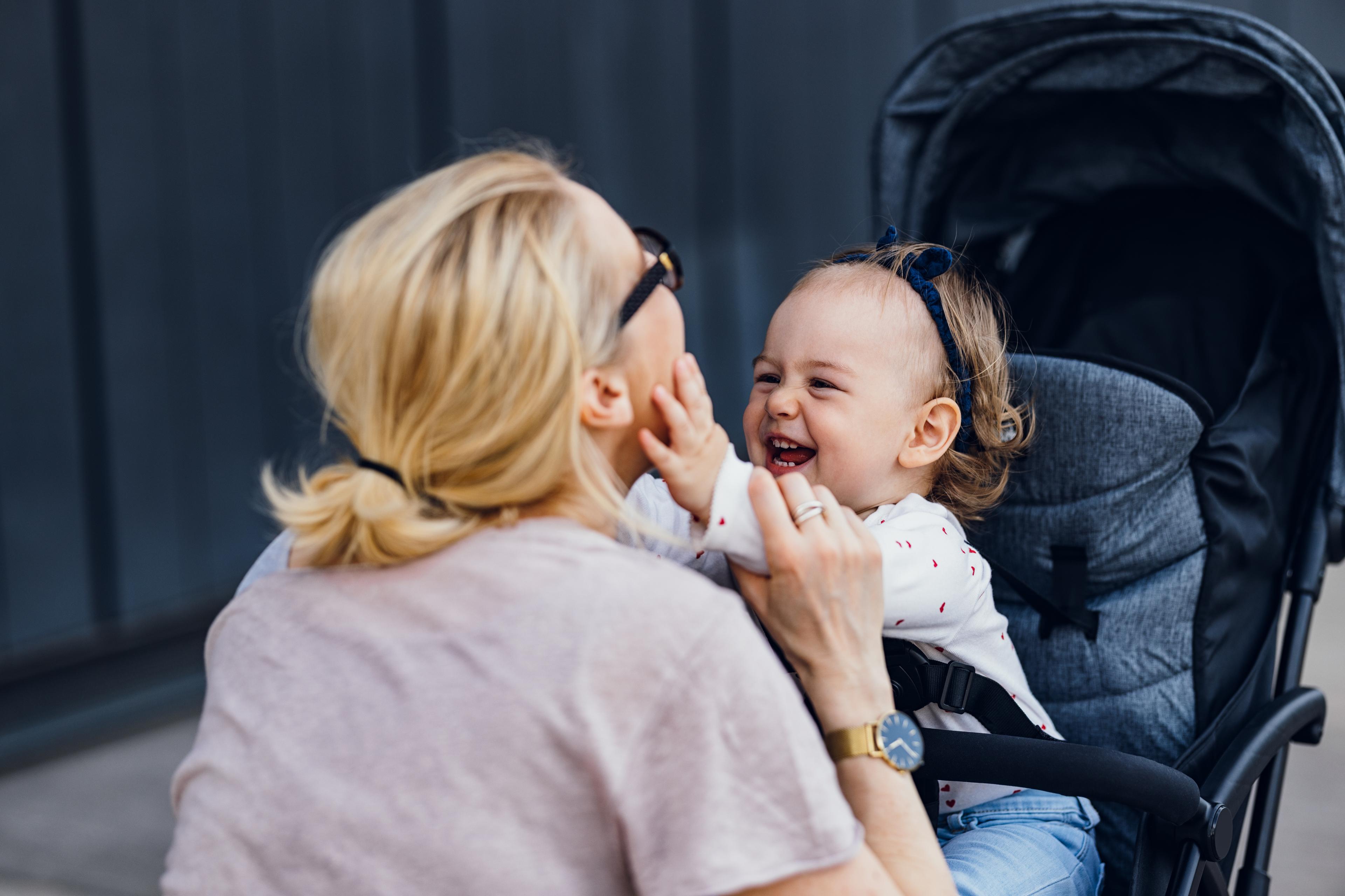Photo of a woman playing with a smiling baby in a pram outside. The woman wears glasses and has her hair tied back.