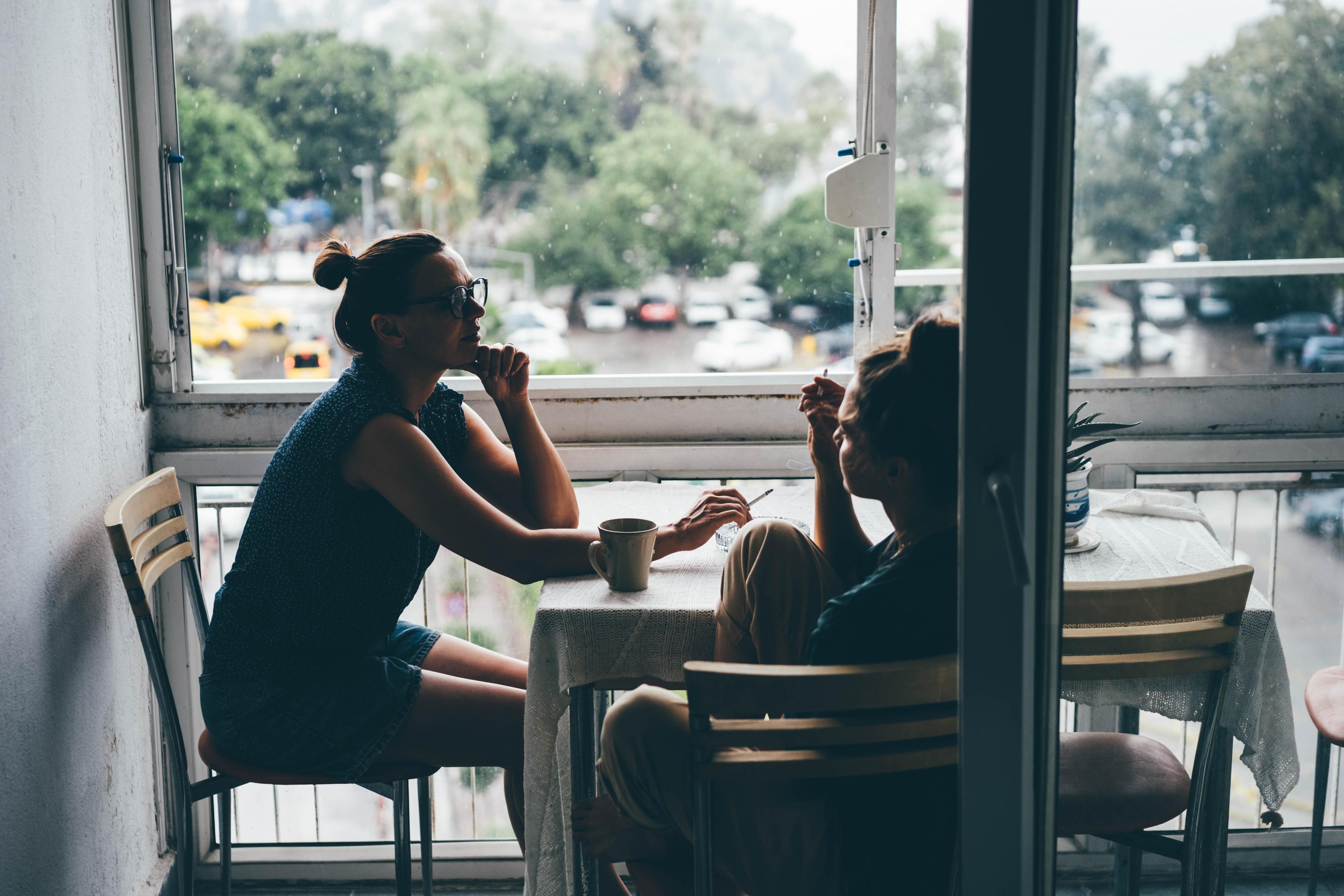 Photo of two people sitting at a table by a window, talking and drinking coffee, with a rainy view outside.