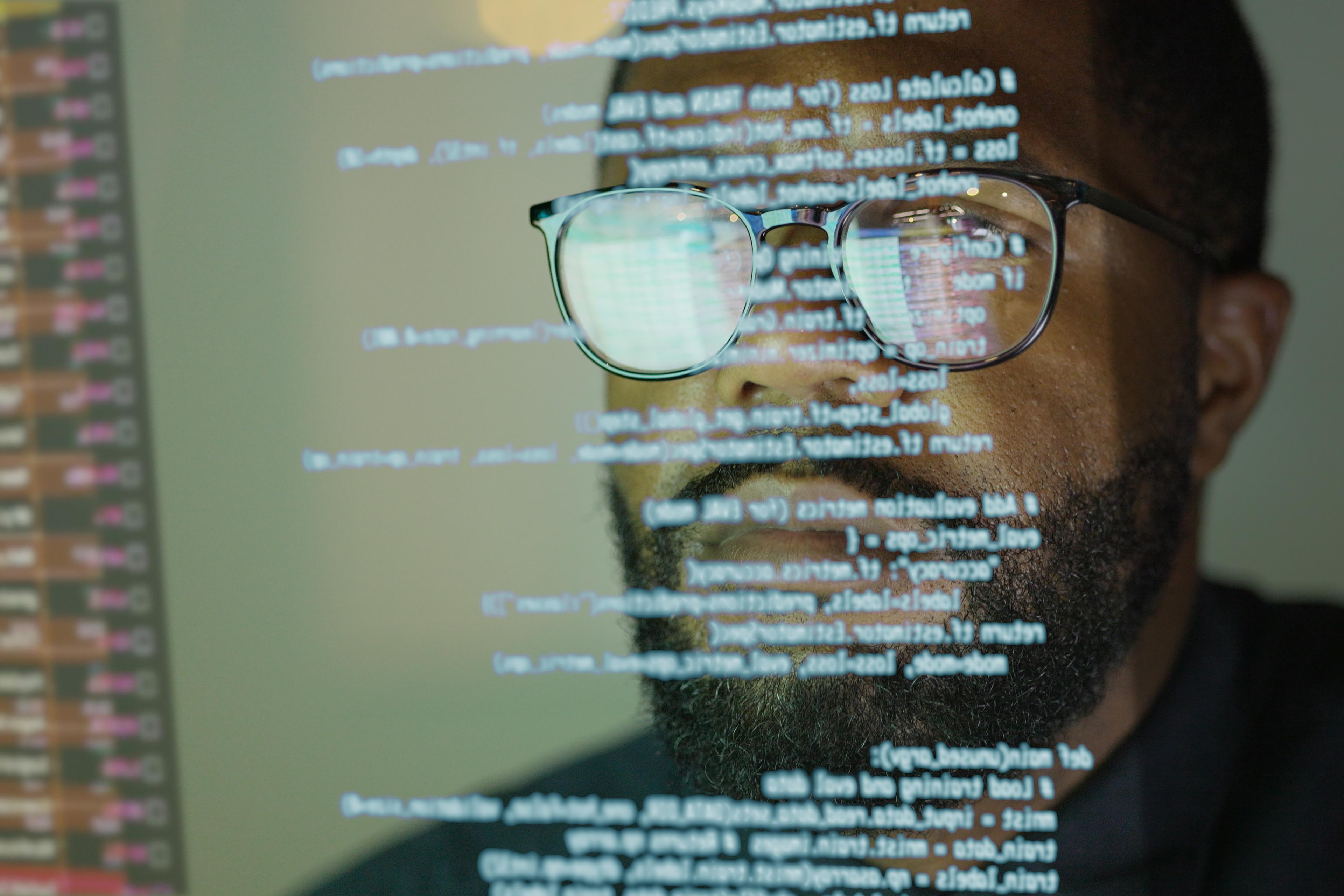 Photo of a person with code reflecting on glasses, focused on a computer screen displaying programming text.