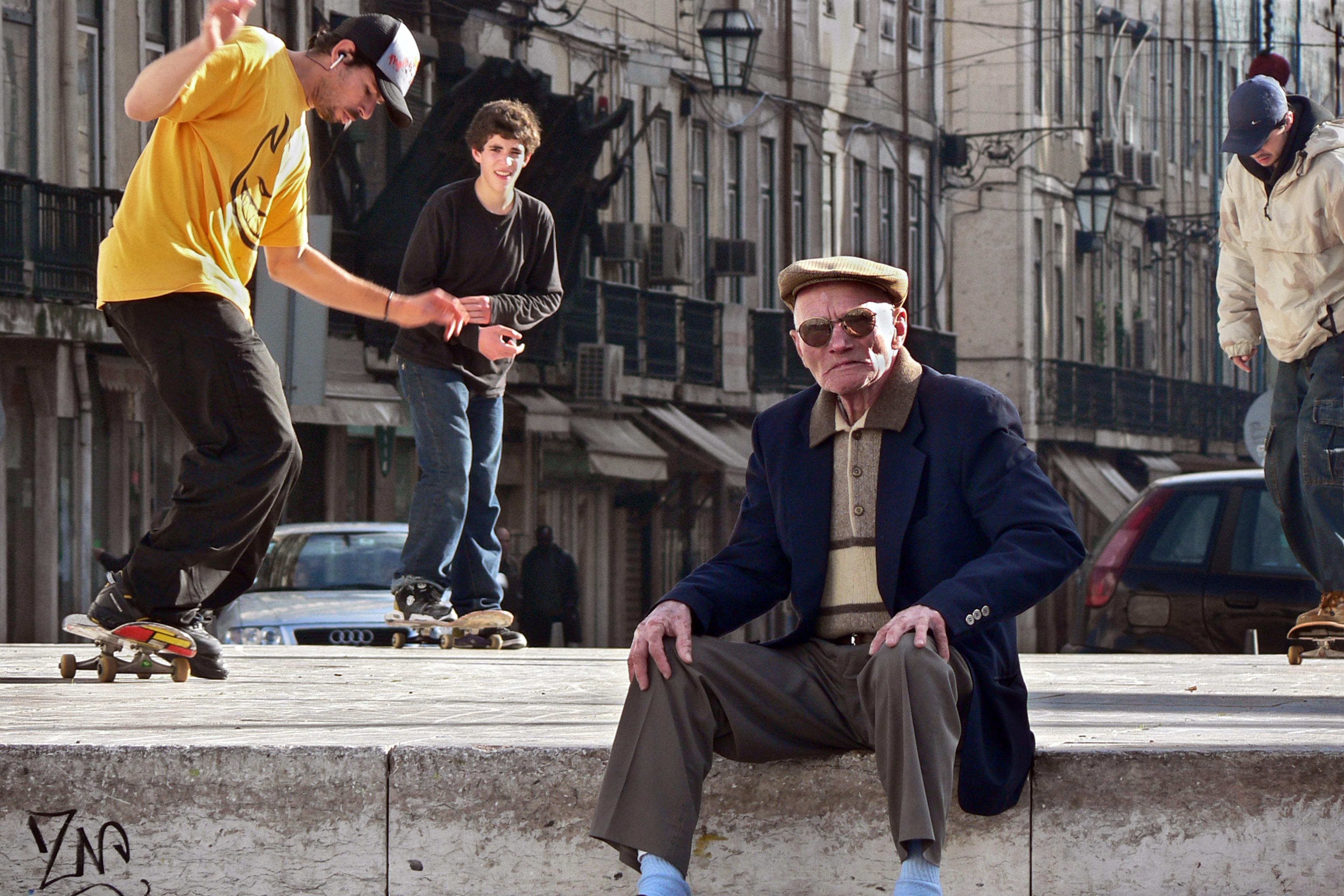 An elderly man seated on a city street as skateboarders perform tricks around him.