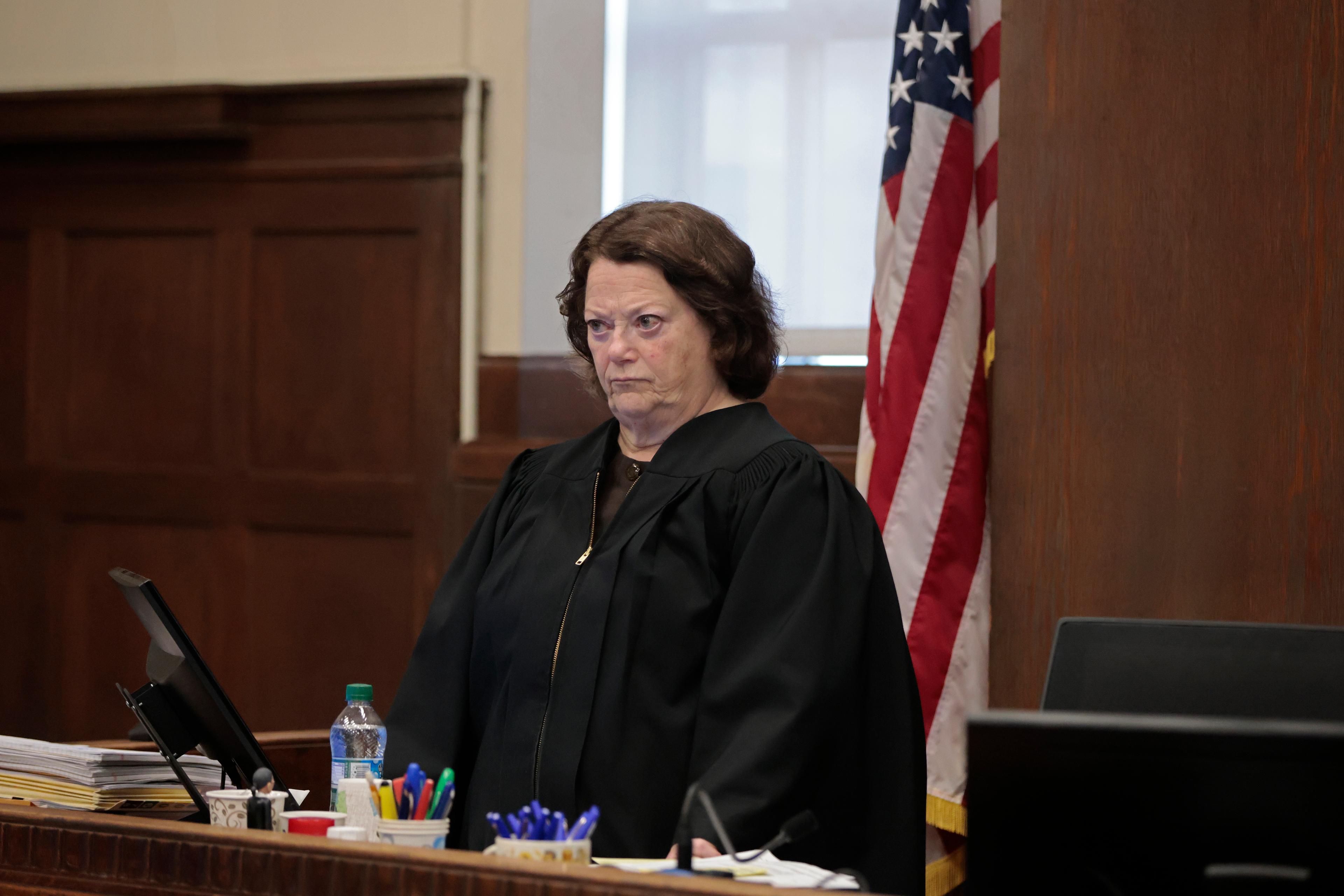 A judge with a stern expression in a courtroom wearing a black robe standing in front of a US flag.