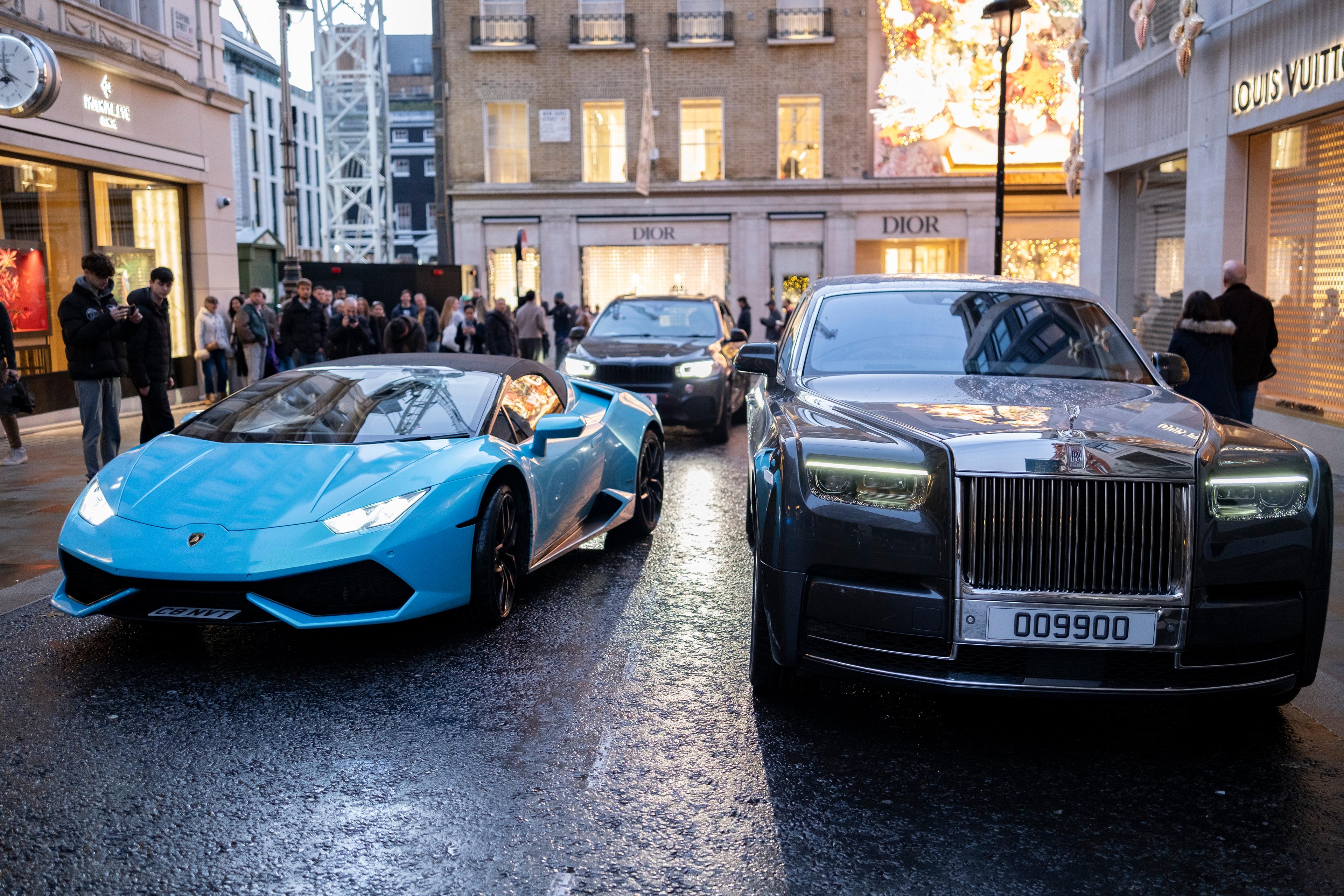 A blue sports car and a grey luxury car parked on a city street with people and a Christian Dior shop in the background.