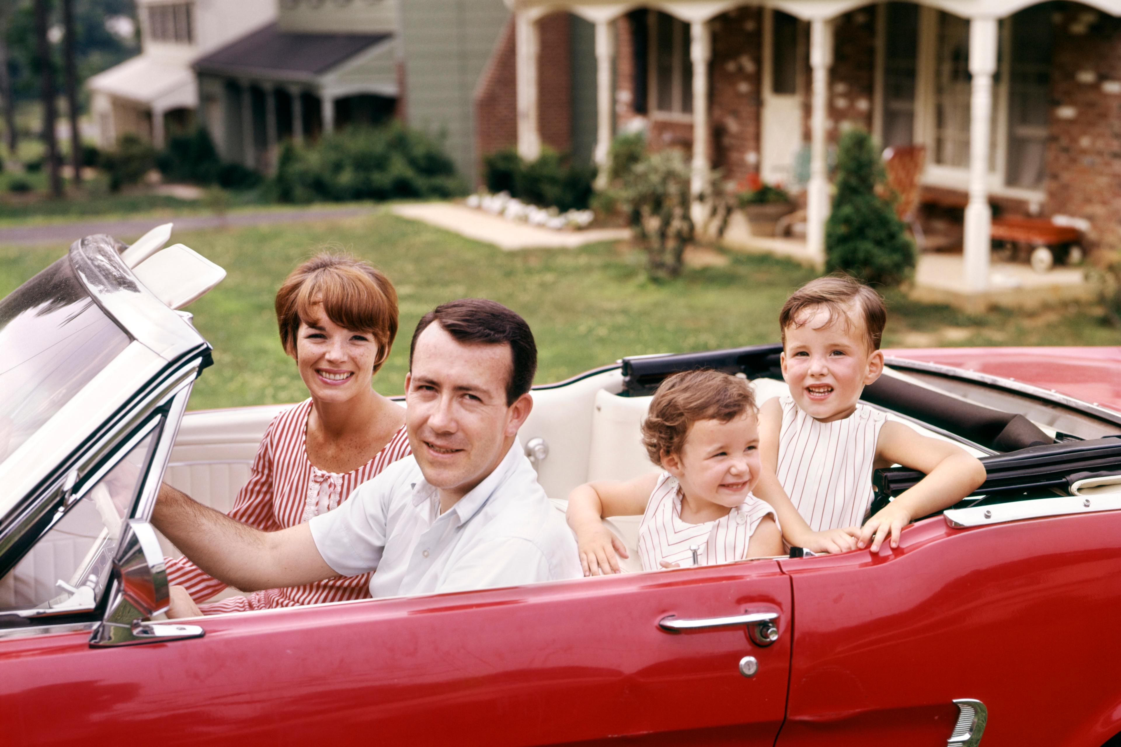 Photo of a smiling family sitting in a red convertible car parked in front of a suburban house.