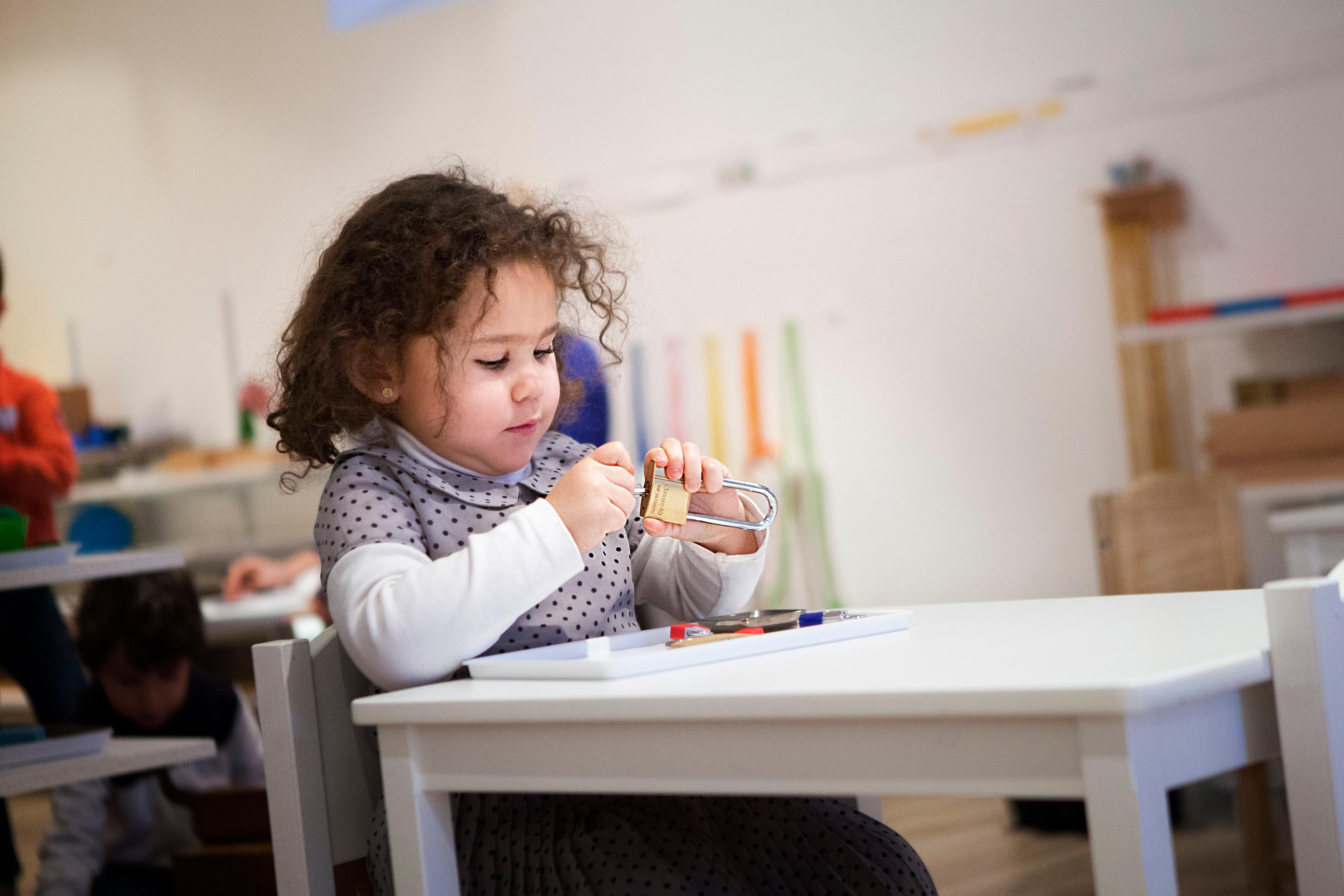 Photo of a young child in a classroom sitting at a table playing with a padlock, with blurred shelves and children in the background.