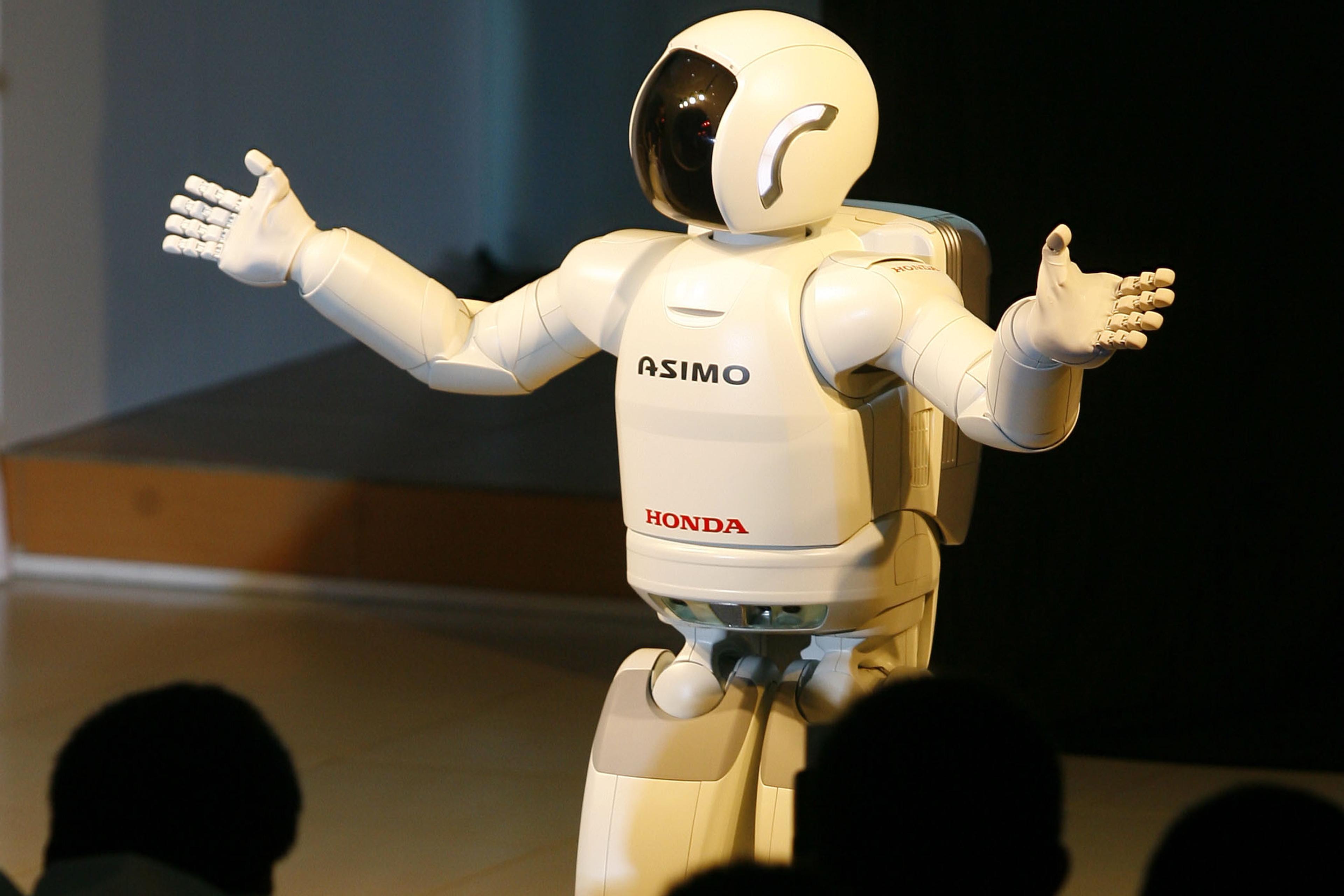 Photo of a humanoid robot with outstretched arms on a stage facing an audience in a dimly lit room.