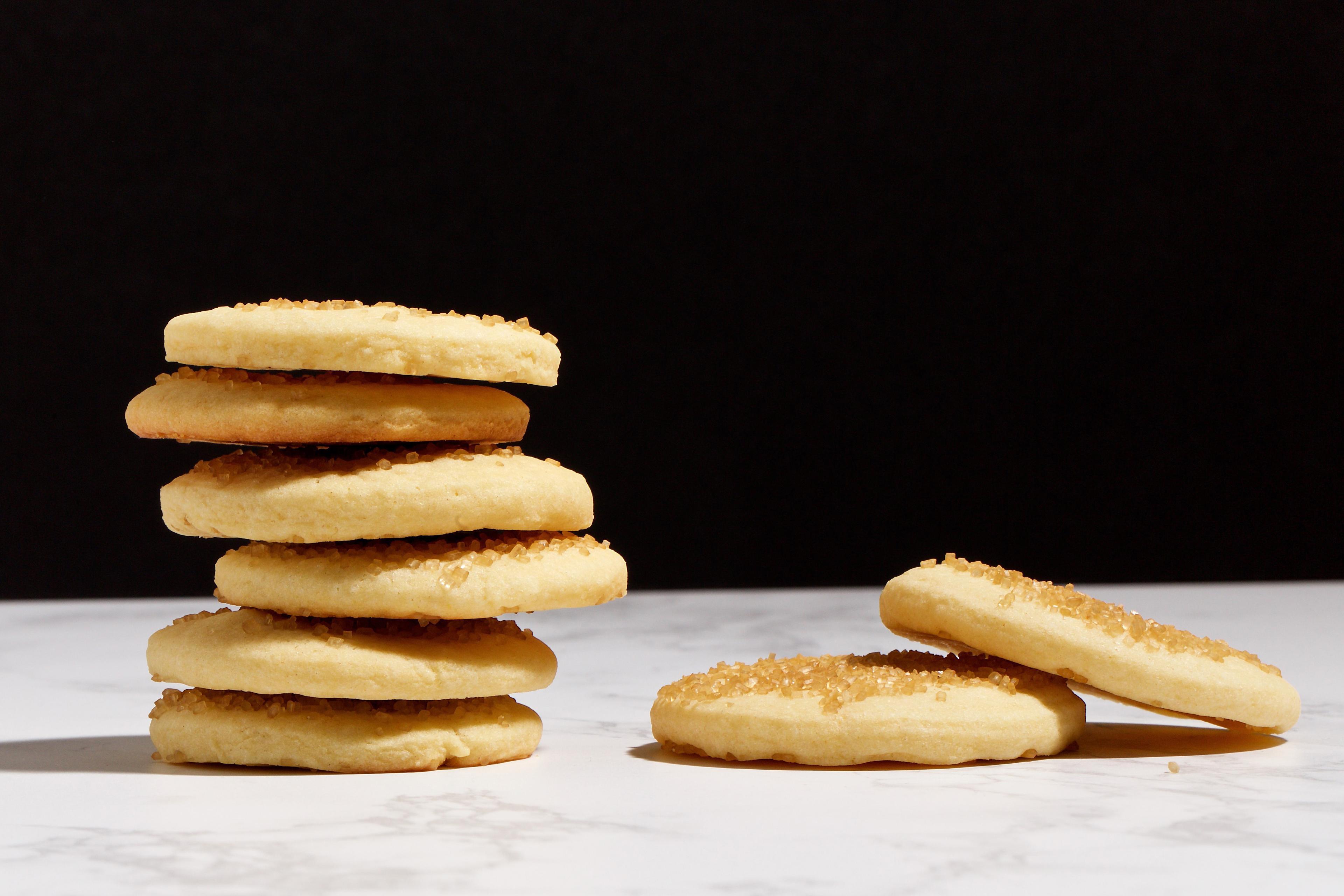 Photo of a stack of six sugar cookies on a marble surface with two cookies lying flat beside them against a dark background.
