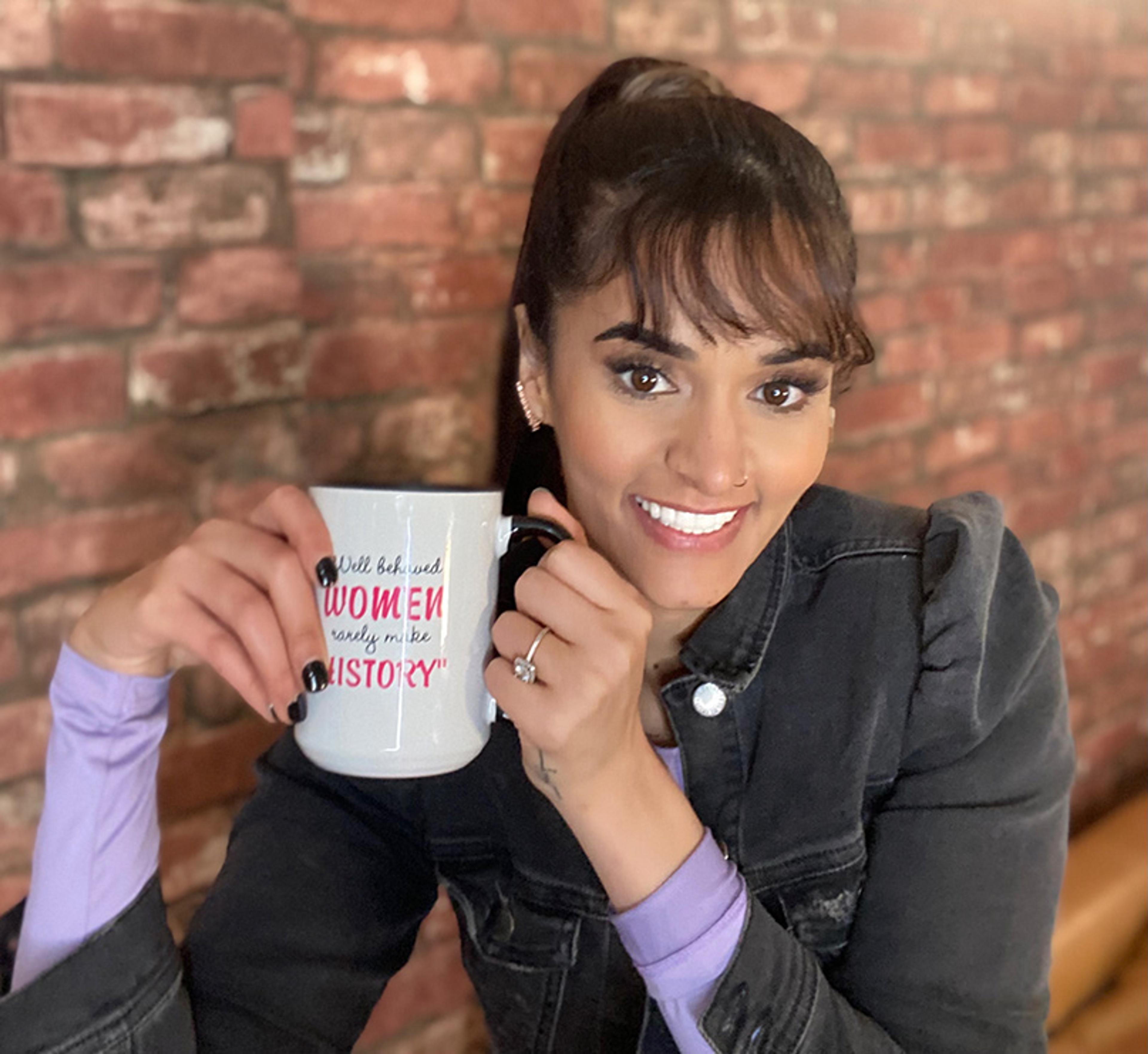 A woman smiling, holding a mug with text ‘Well behaved women rarely make history’, against a brick background.