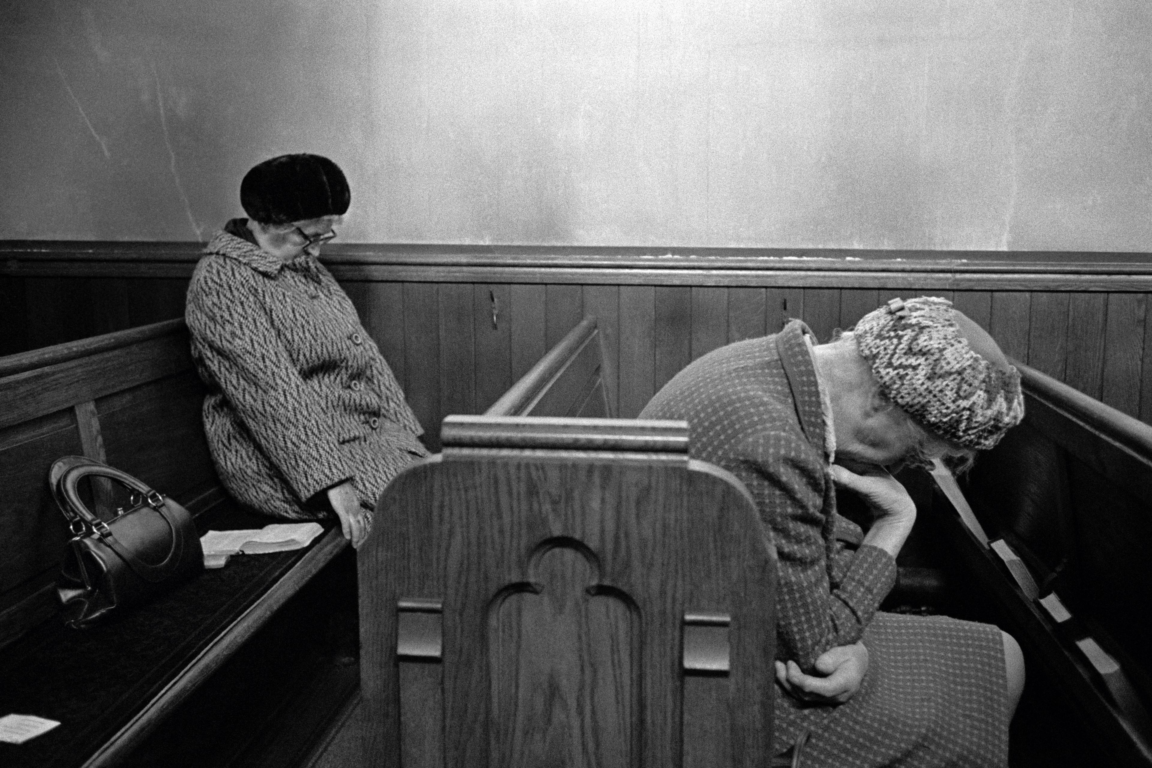 Black and white photo of two elderly women sitting on a wooden bench in a chapel with heads bowed, one has a handbag beside her.