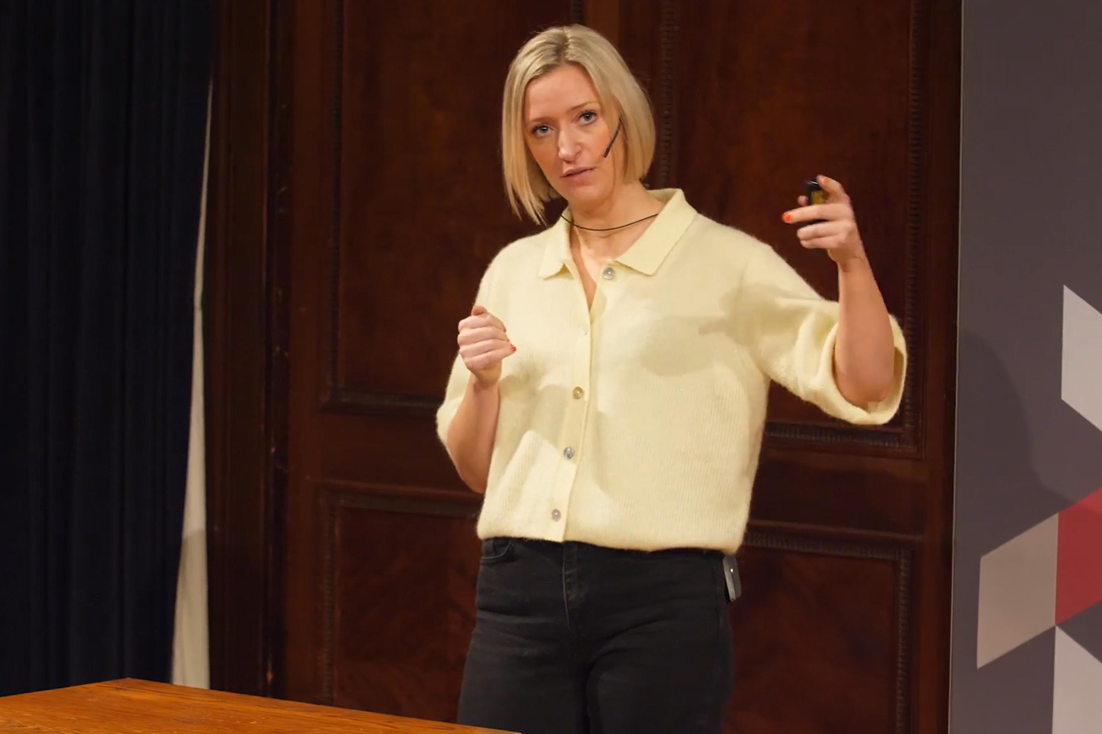 A woman with a headset giving a presentation. She gestures with her hand. The background is wood paneling.