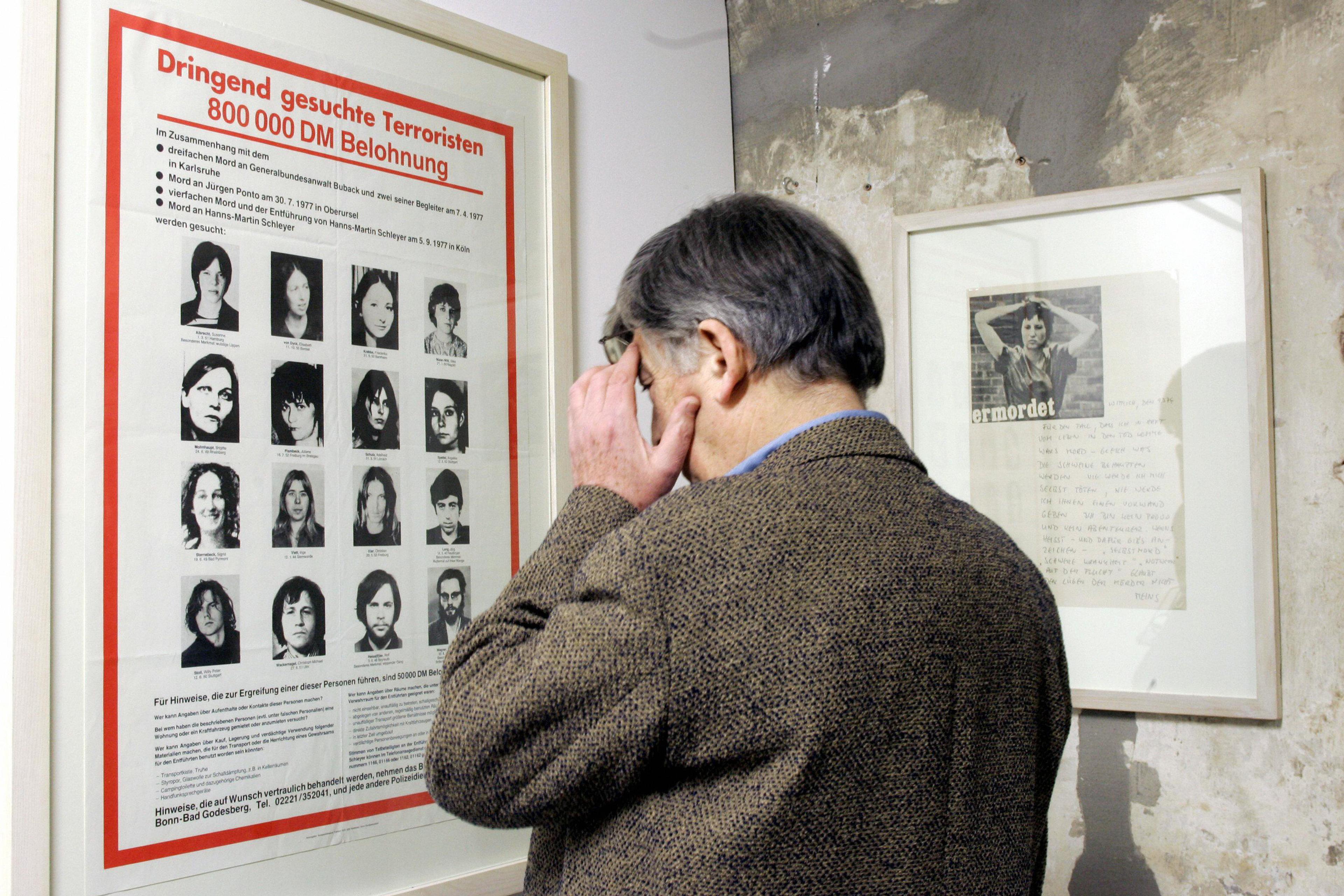 A man in a tweed jacket viewing a framed German wanted poster on a wall in a museum or gallery setting.