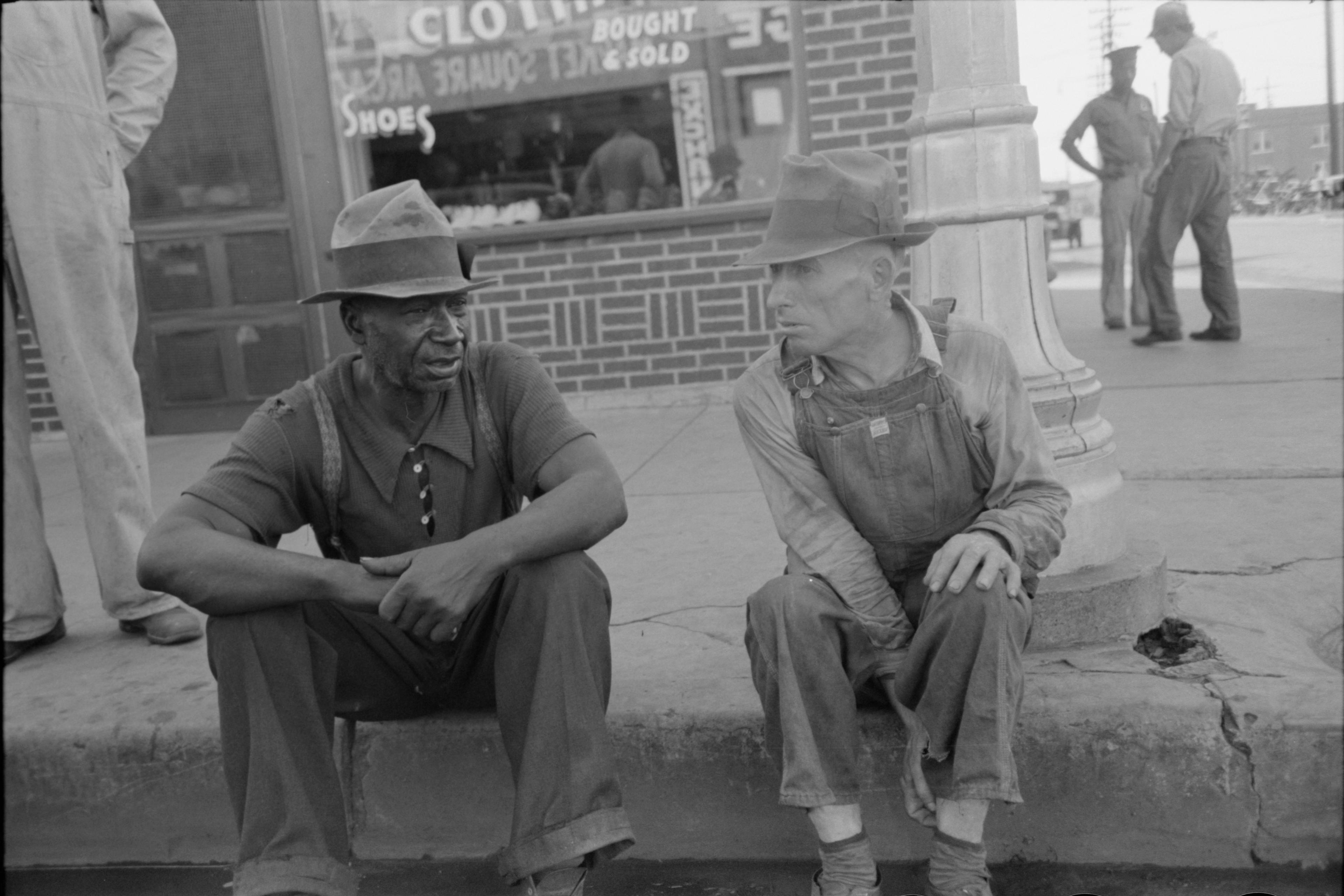 Black and white photo of two men sitting on a kerb in conversation with a brick storefront behind them.