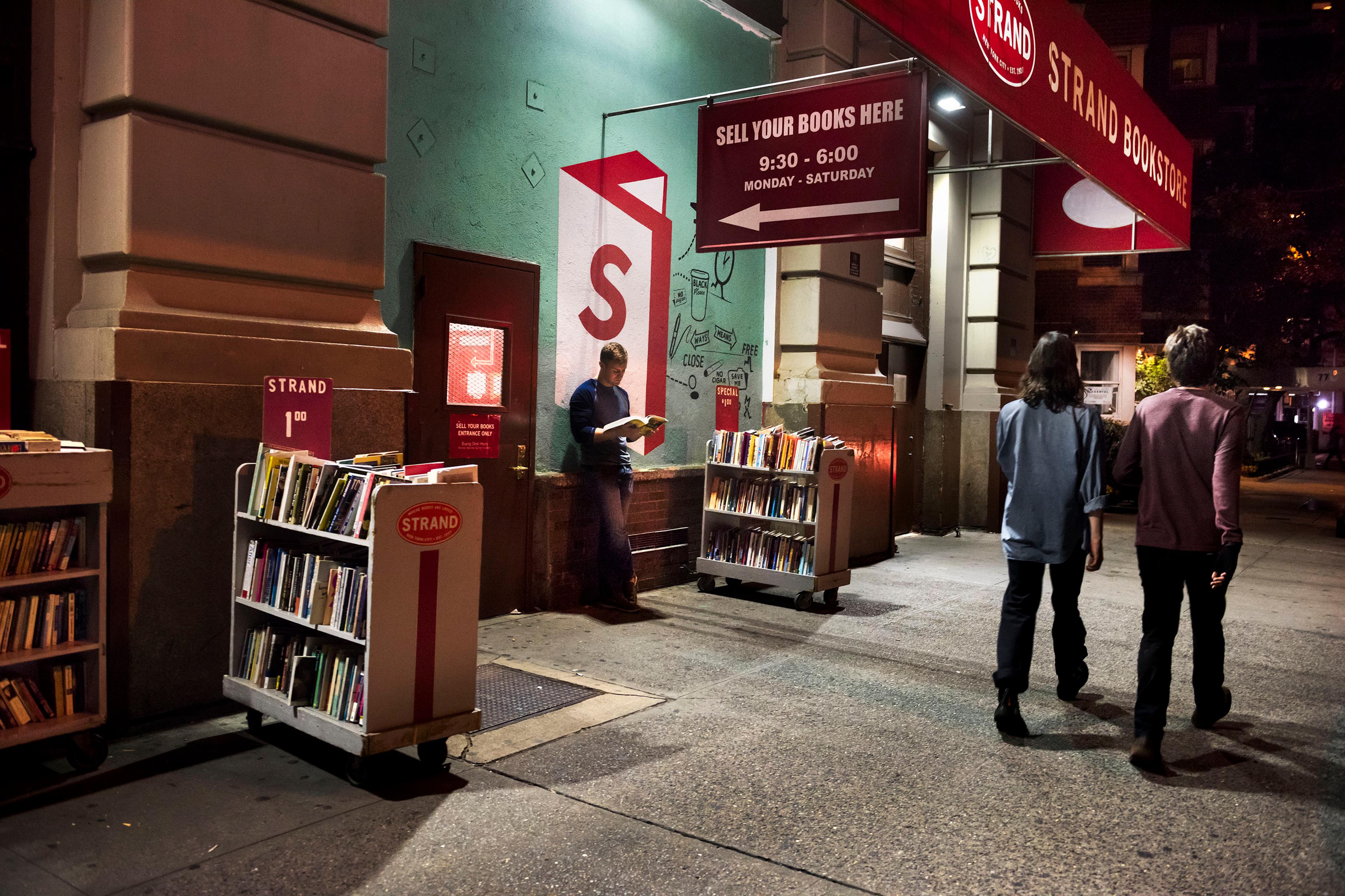 Photo of a bookstore entrance at night with people walking by and a person reading among book trolleys on the pavement.