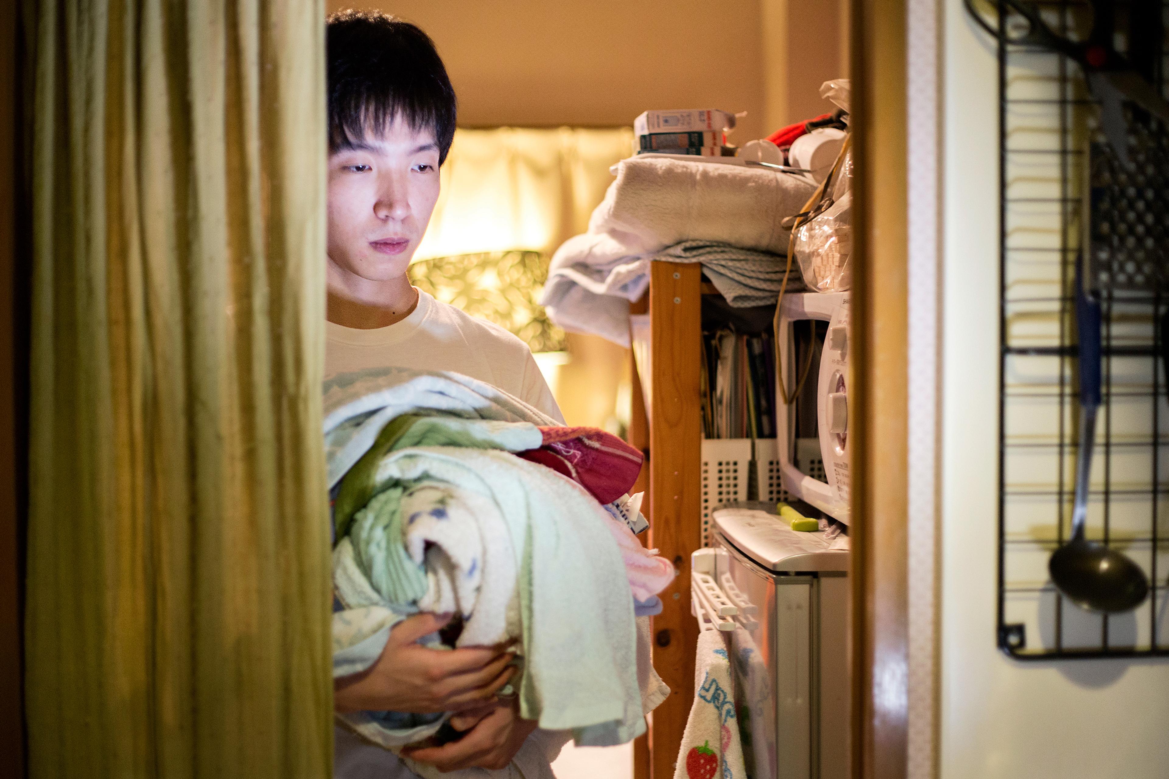 Photo of a man holding laundry in a dimly lit room with shelves, a microwave and a curtain on the left.