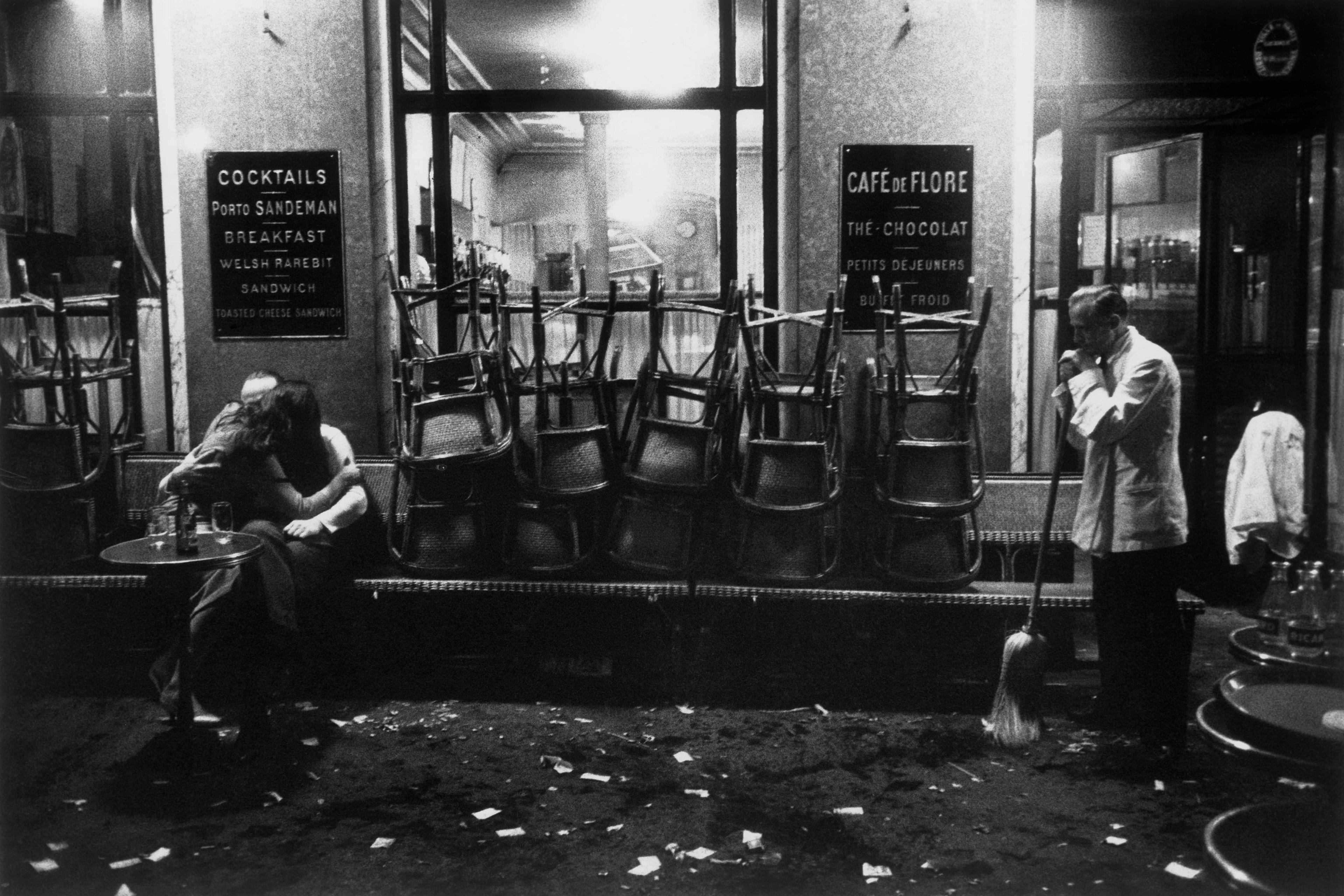 Black and white photo of a couple kissing outside a café at night with a waiter sweeping the floor.