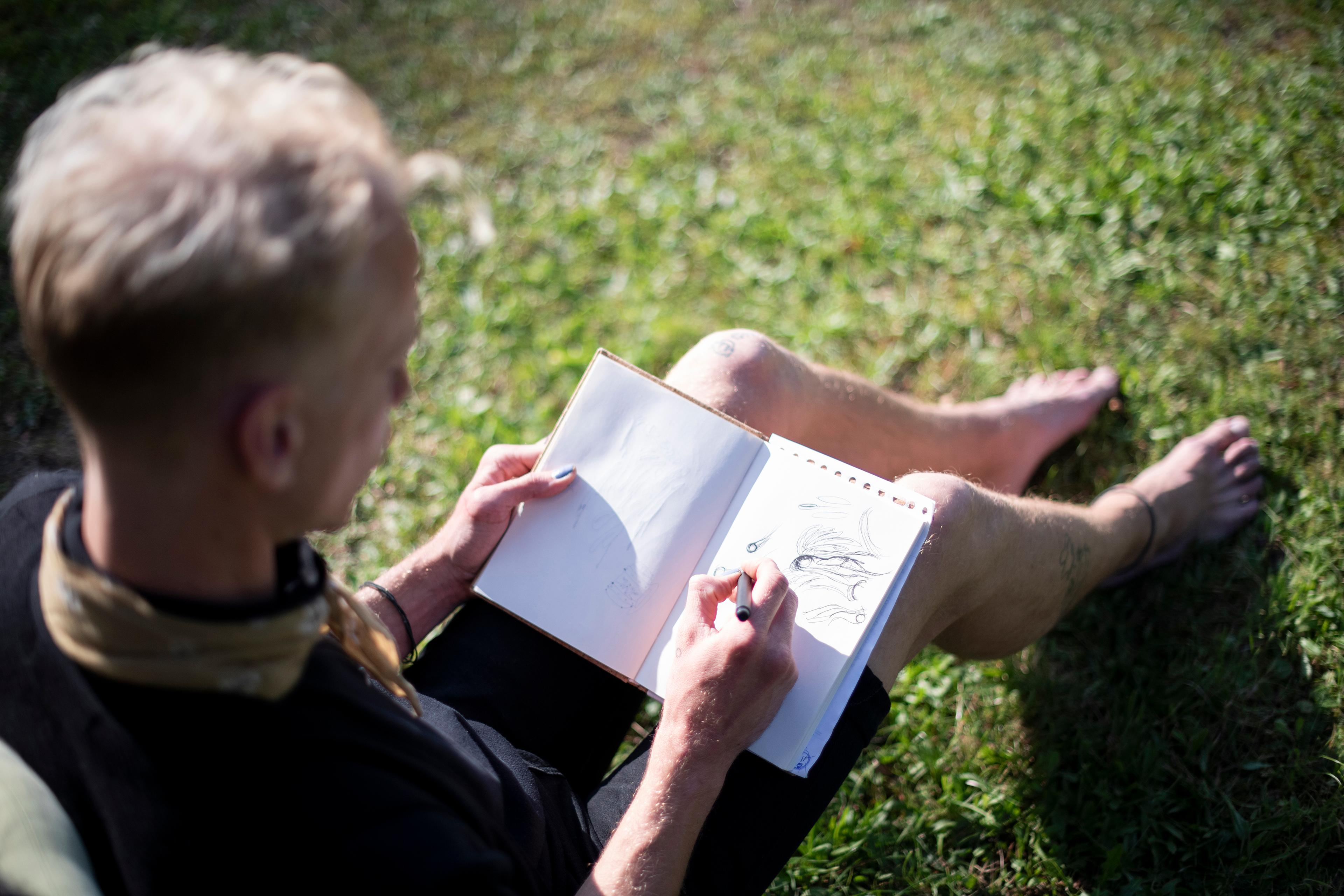 Photo of a person sitting on grass sketching in a notebook, showing their hands and feet in a relaxed outdoor setting.