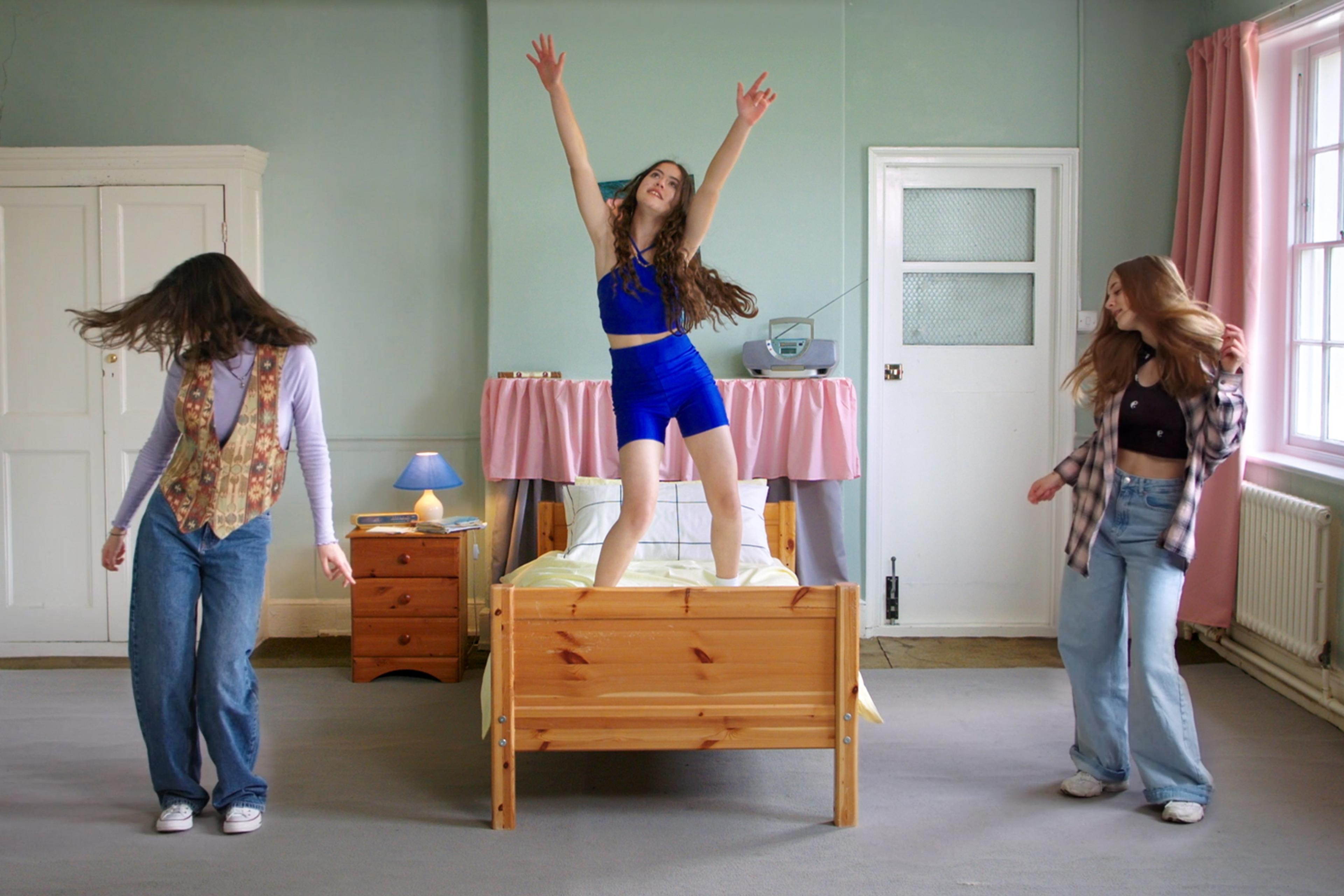 Three girls dancing in a bedroom; one on a bed in blue outfit, others on the floor wearing casual clothes. The room has pink curtains and a blue lamp.