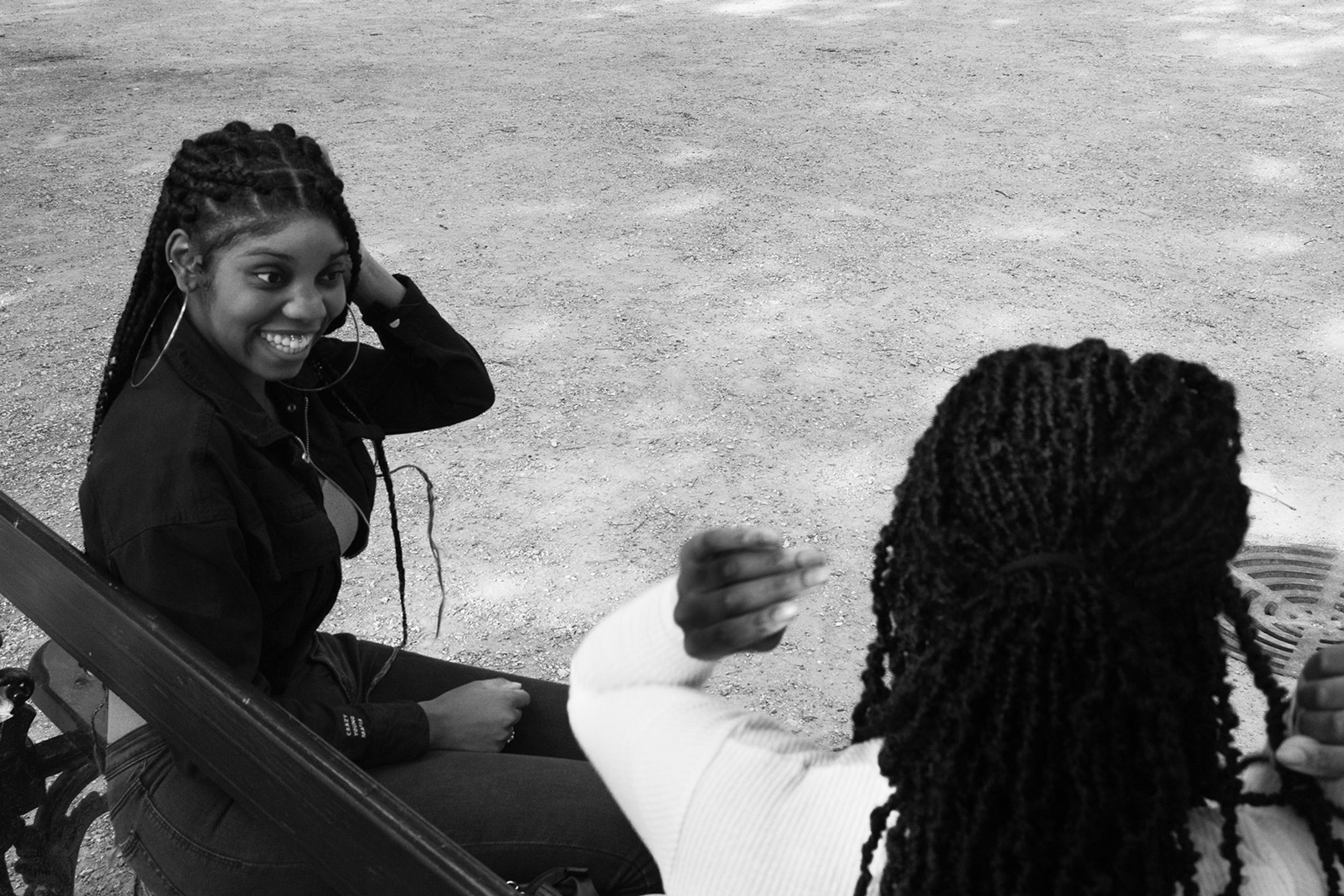 Two women with braided hair sitting on a park bench, smiling and engaging in conversation. One faces the camera, while the other is seen from behind.