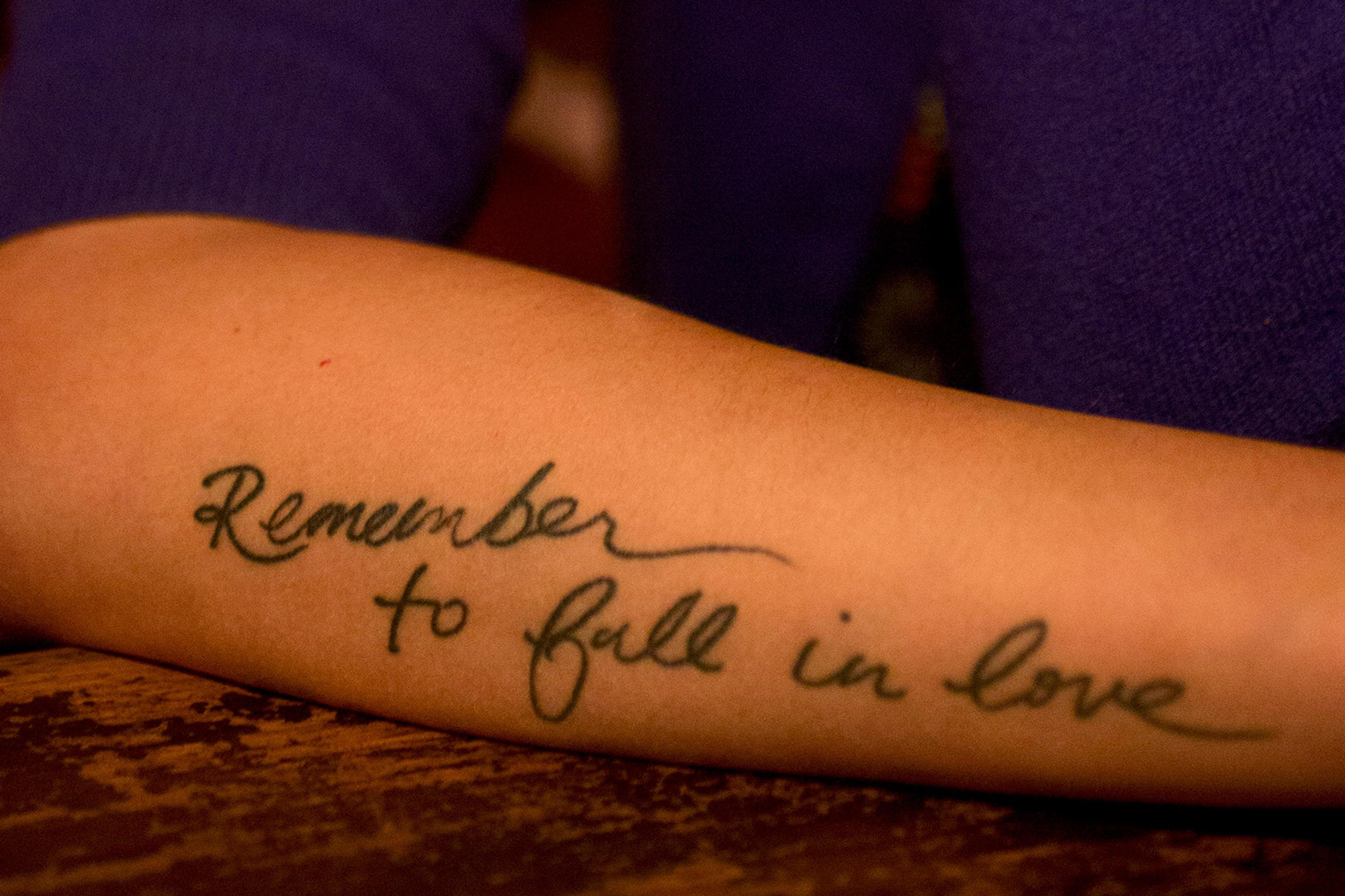 Forearm tattoo in cursive script reads “Remember to fall in love” in black ink.