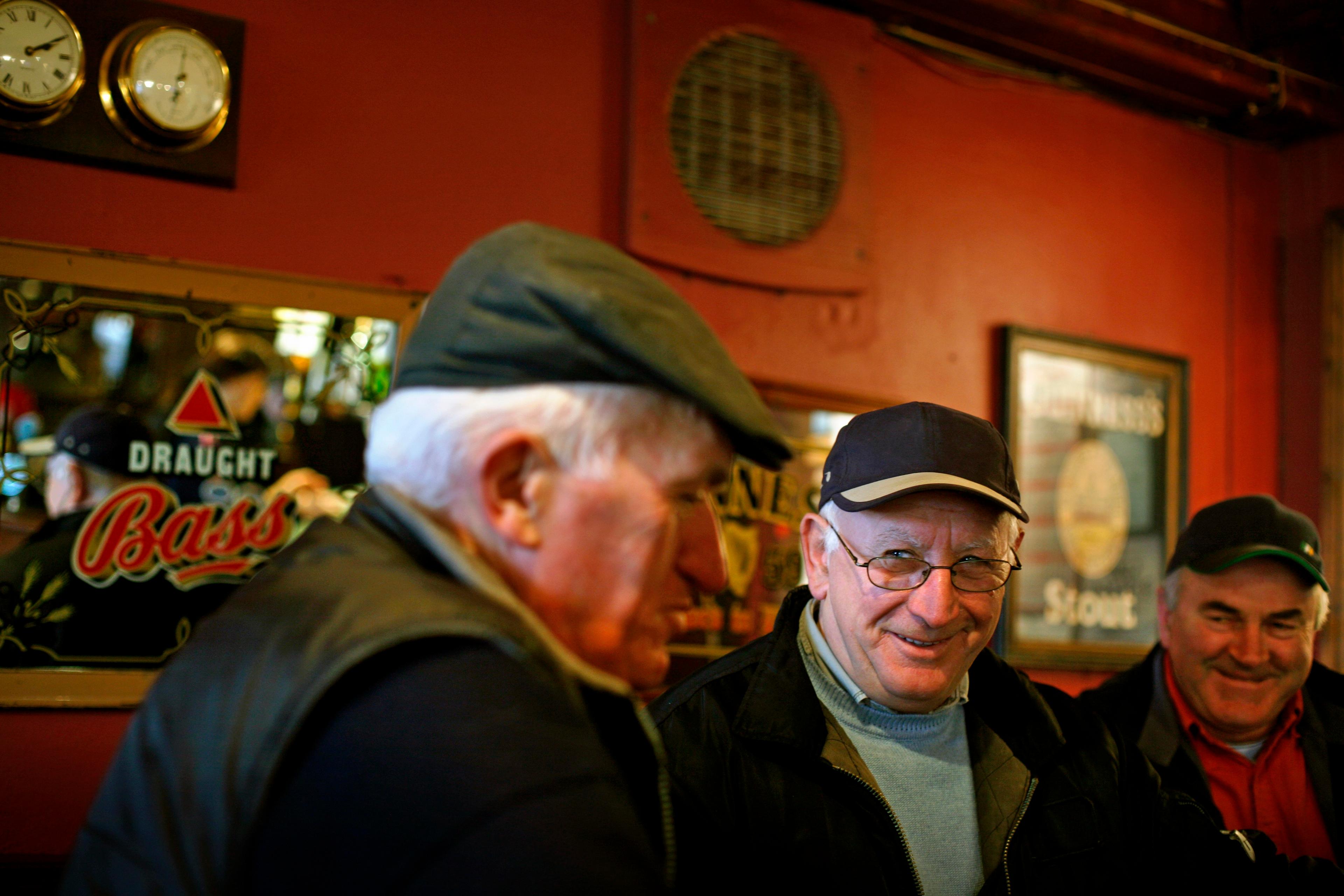 Photo of three older men in a pub, smiling and wearing caps. The background has pub mirrors and a red wall.