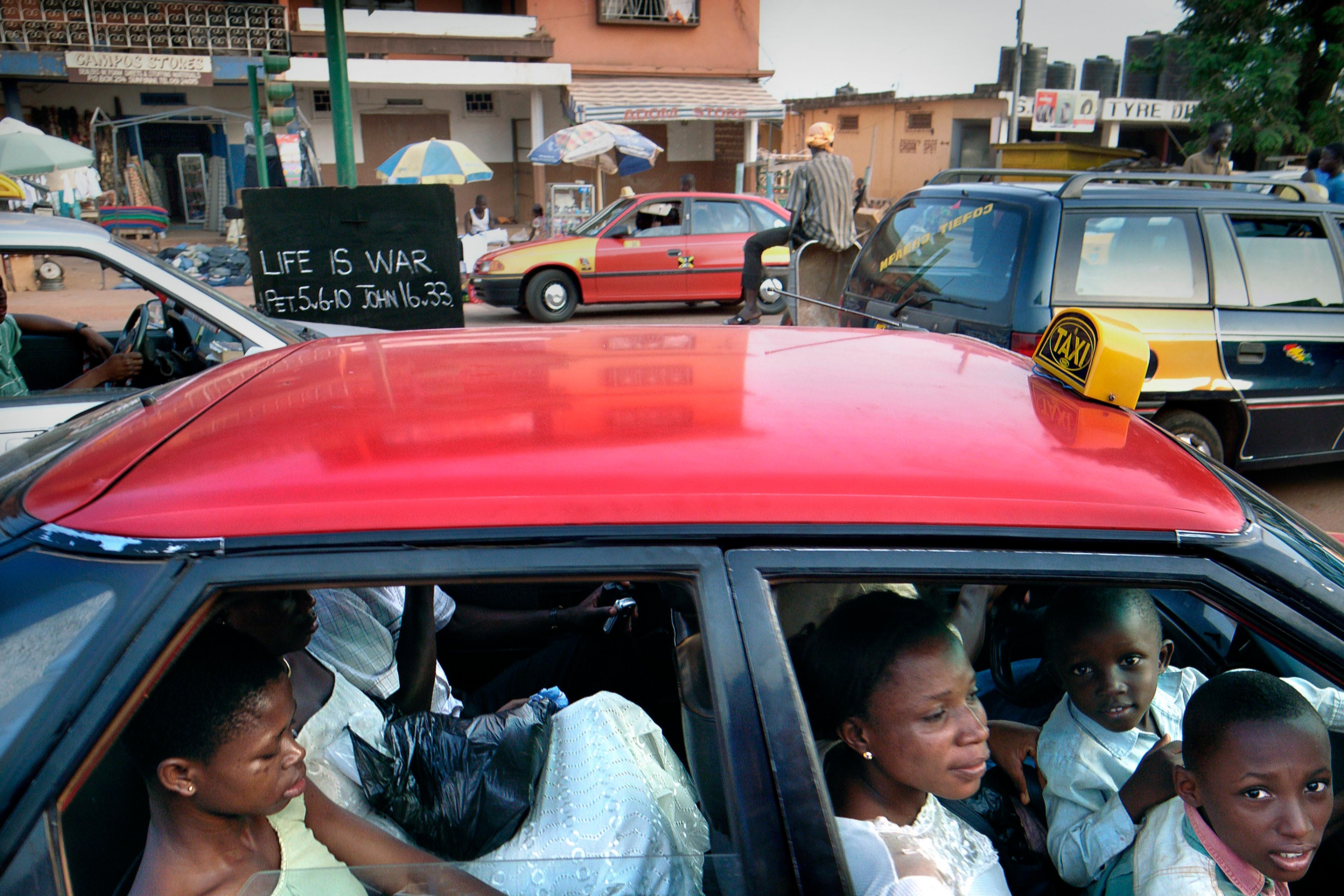 Photo of a red taxi with passengers in an urban setting, a sign saying “Life is War” visible in the background.