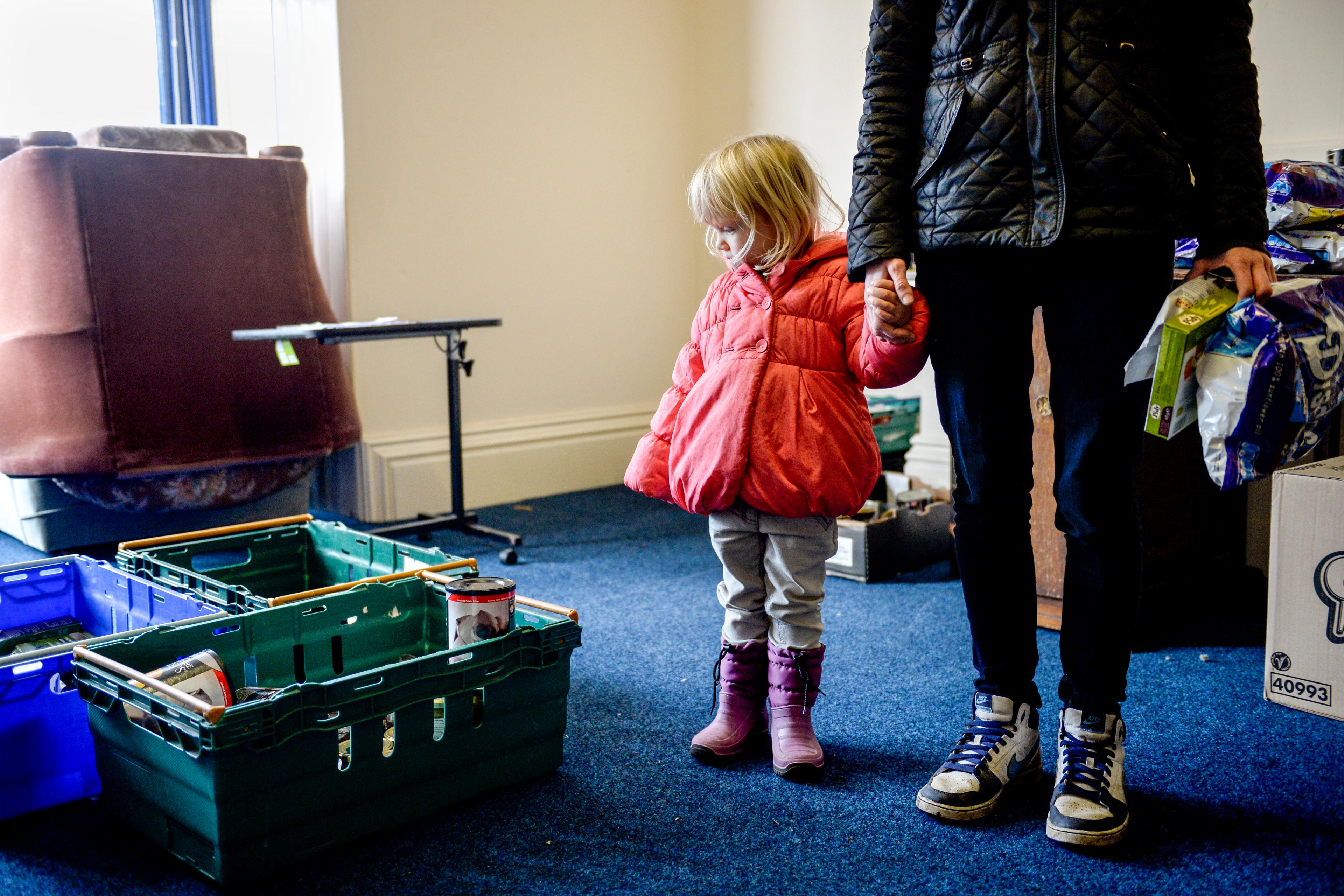 Photo of a child in a pink coat holding an adult’s hand in a room with crates containing canned goods.